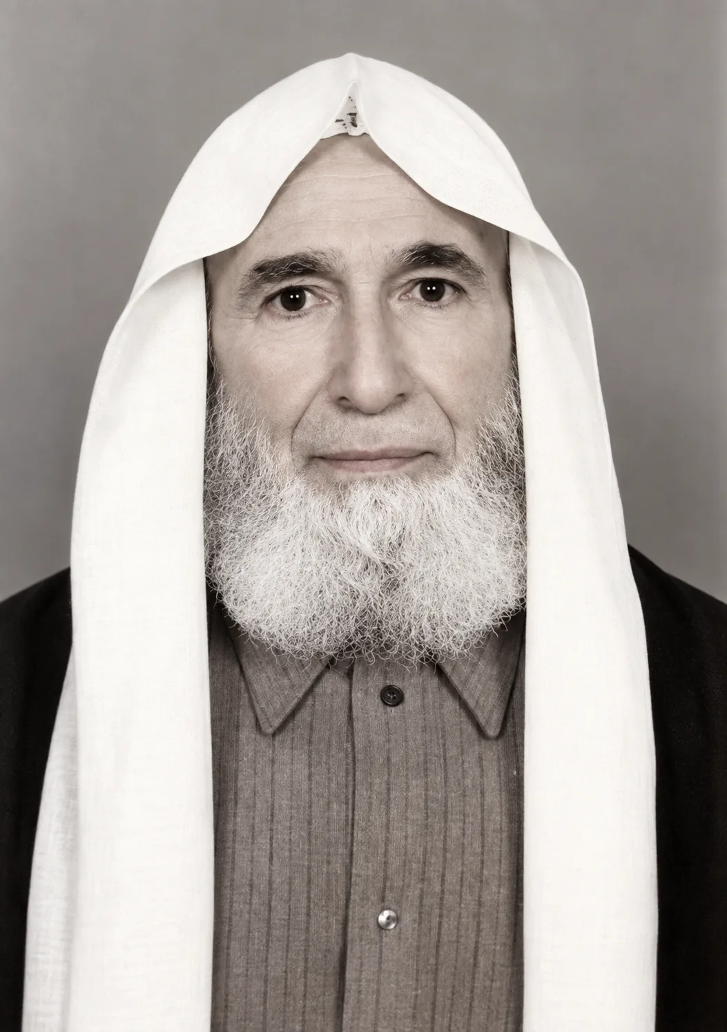 Formal frontal portrait in near-monochrome of a man with a grey and white beard, wearing a white headcloth draped over a cap, a dark robe over a collared shirt, against a plain grey background
