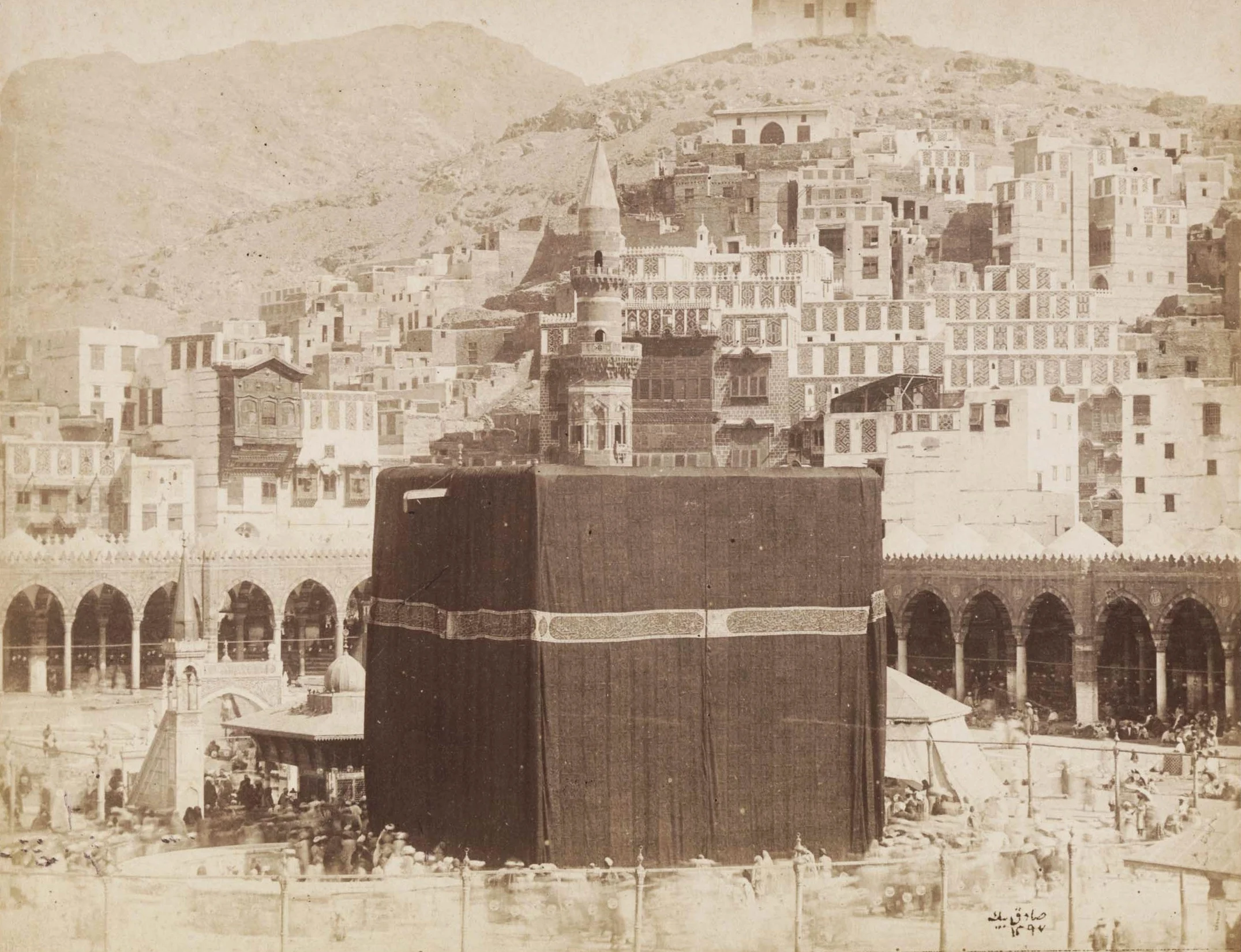 A sepia photograph of the Kaaba draped in its dark kiswah, seen from above the mosque courtyard with the surrounding colonnade and the multi-storeyed buildings of old Mecca rising up the hillside behind it, pilgrims gathered at its base