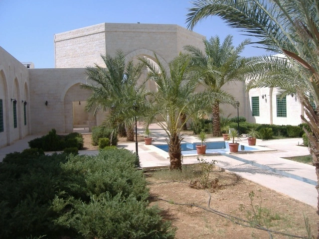 A stone mausoleum building with arched arcades set behind a garden courtyard planted with date palms and shrubs, with a small pool along the paved walkway