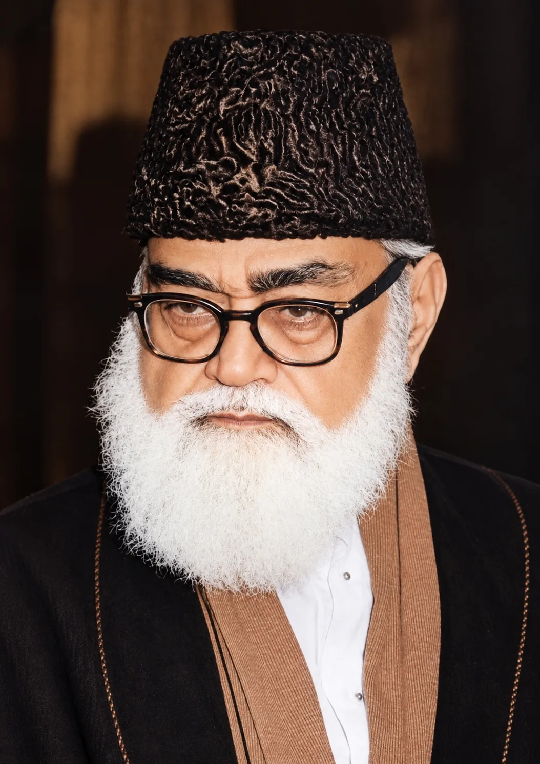 AI-generated impression: Close portrait of an elderly man with a full white beard, wearing a tall black astrakhan cap, heavy dark-rimmed spectacles, a white shirt and a tan shawl over a dark jacket, against a dark background