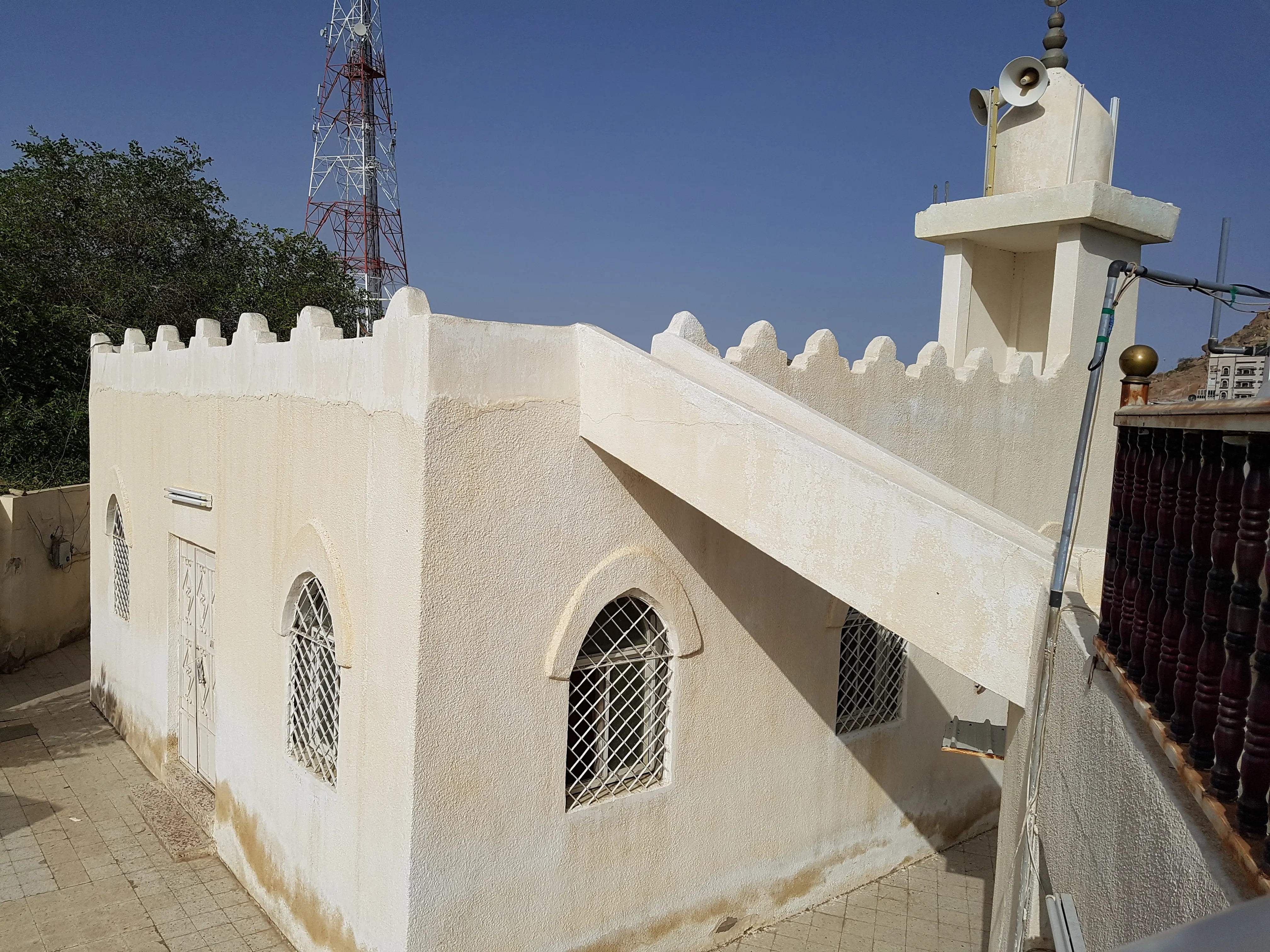 A small whitewashed mosque with a crenellated parapet, arched windows set with white grilles, an external stair rising to the roof, and a square minaret carrying loudspeakers, against a clear sky