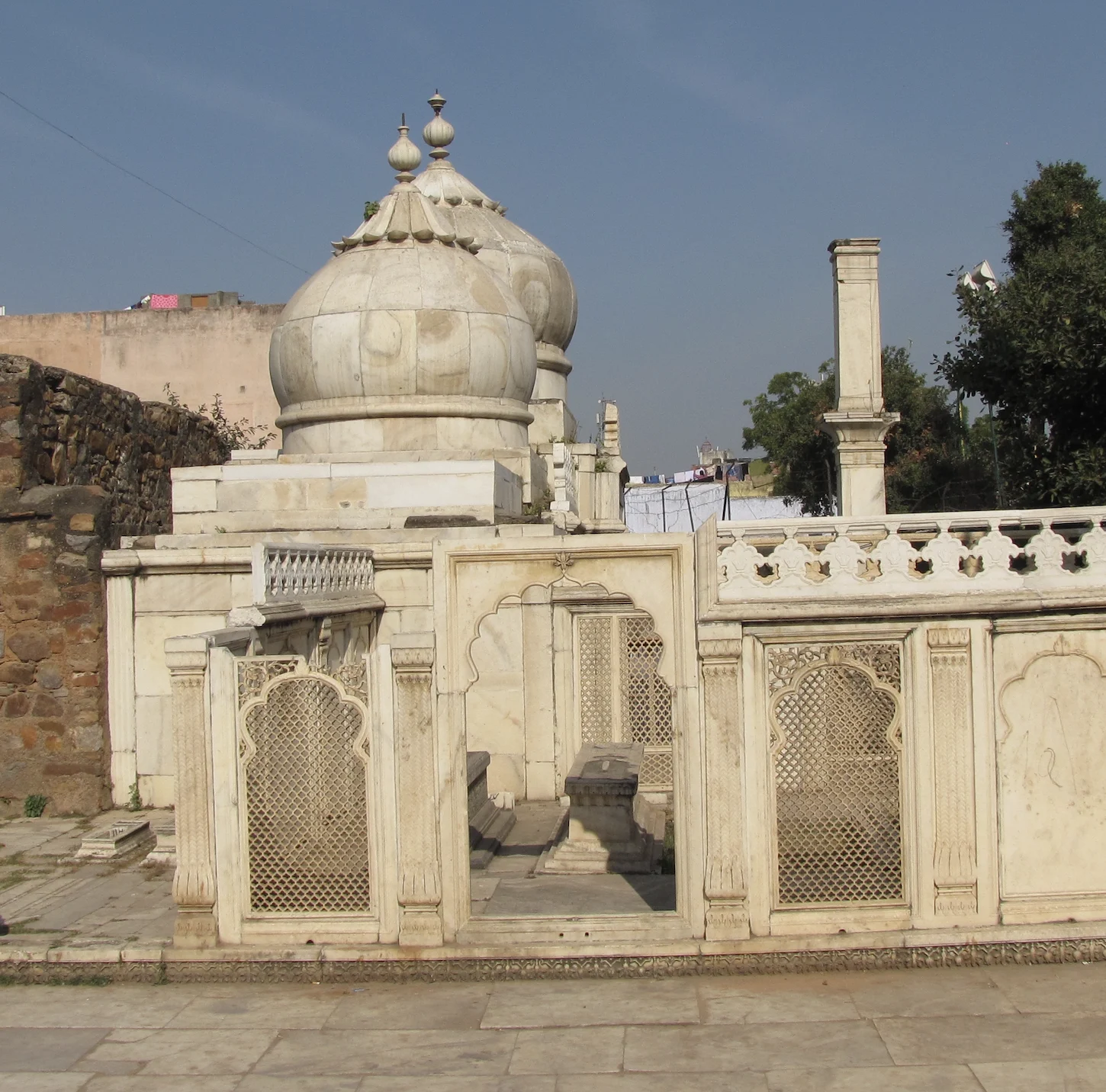 Two white marble domed tombs enclosed by a carved marble screen with pierced lattice panels and arched niches, set against an old stone wall and a tree, in the Zafar Mahal complex at Mehrauli, Delhi