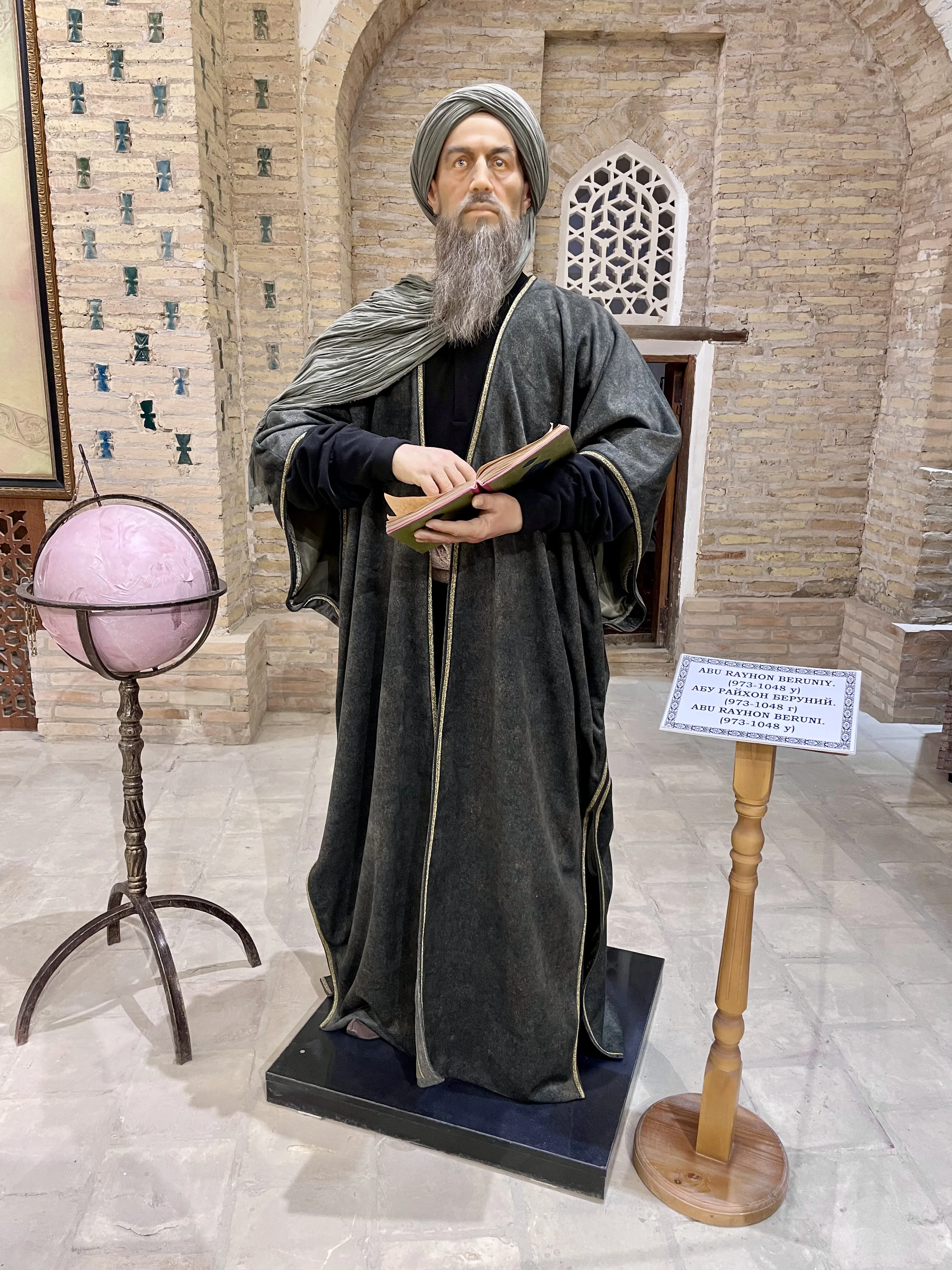 A life-size costumed figure of a bearded scholar in a grey turban and shawl over a dark grey robe, holding an open book, standing on a low plinth in a brick-vaulted hall beside a globe in a metal mounting on an iron tripod and a wooden lectern carrying a name label in three scripts