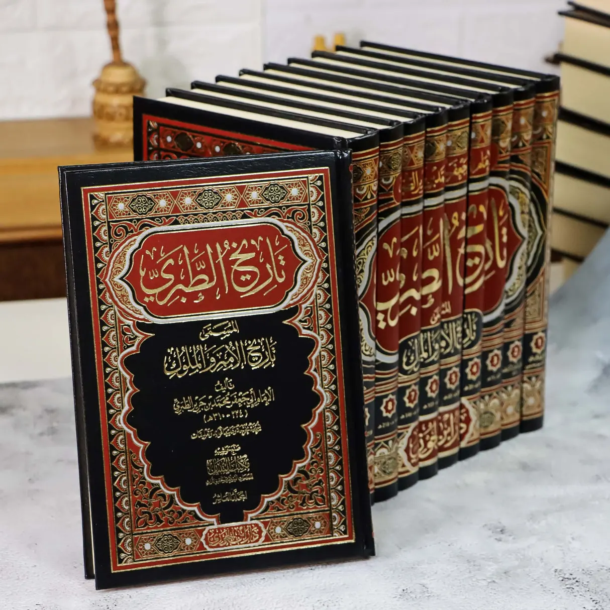 A modern printed edition of Tarikh al-Tabari, the front volume bound in black and red with gilt tooling and its title in Arabic, with the matching volumes of the set ranged behind it