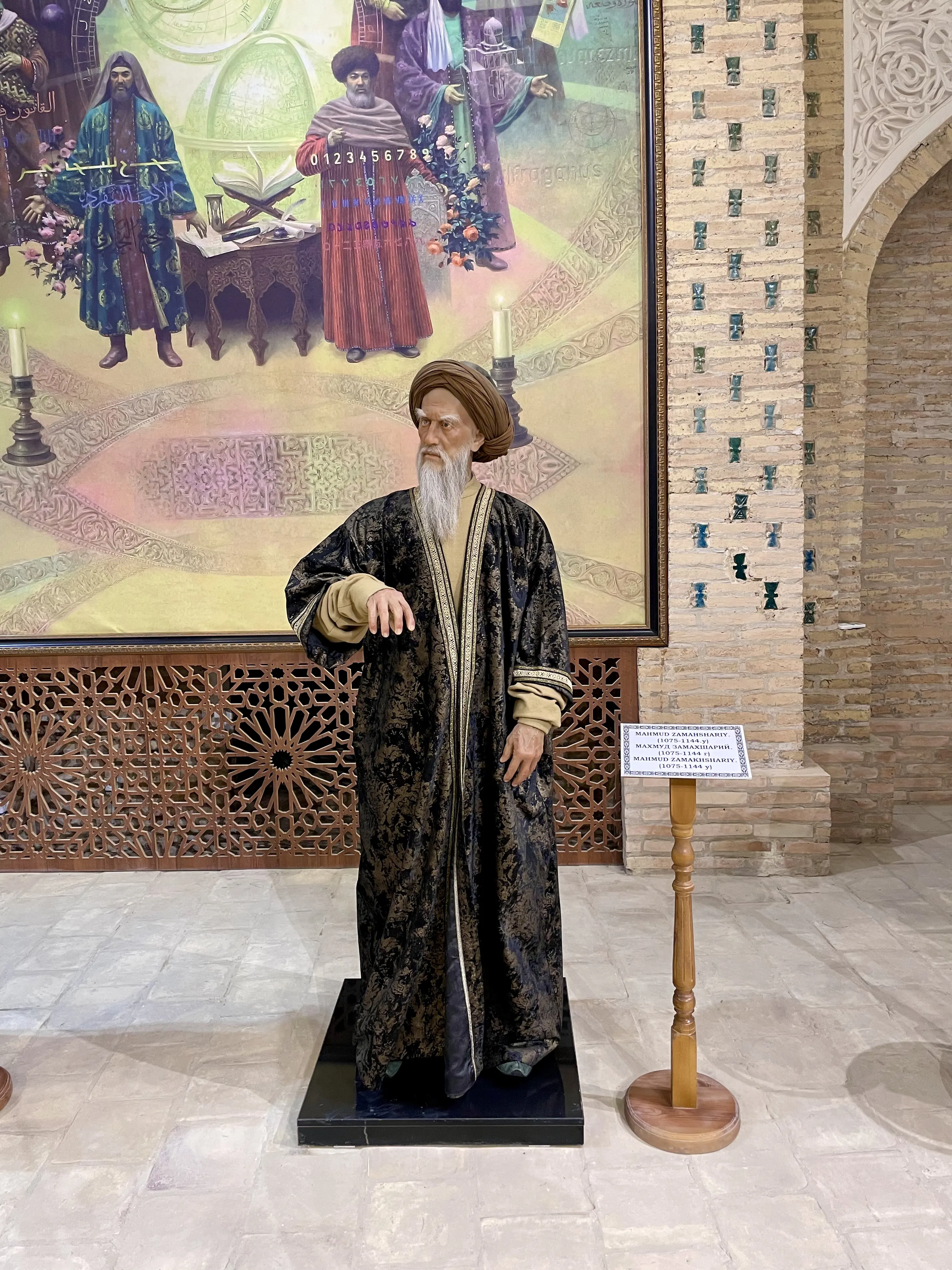 A life-size costumed figure of an elderly bearded scholar in a brown turban and a dark robe patterned with gold brocade over a tan inner garment, one hand extended before him, standing on a low black plinth in a brick-walled hall; behind him hangs a large framed painting of turbaned scholars with books, a globe and a panel of numerals, and beside him a wooden lectern carries a name label in three scripts