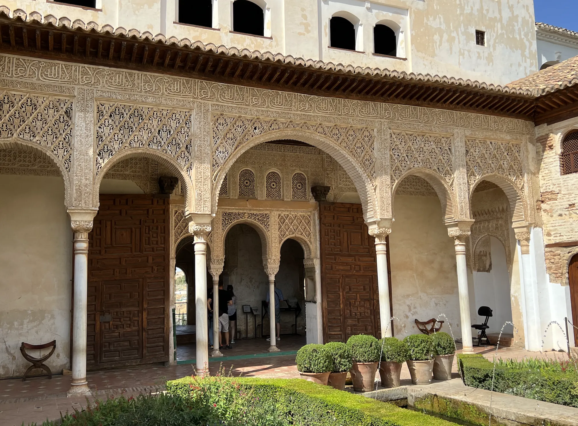 An arcade at the Alhambra with intricately carved stucco arches bearing Arabic inscription, resting on slender white marble columns, beside a garden with a fountain and clipped hedges