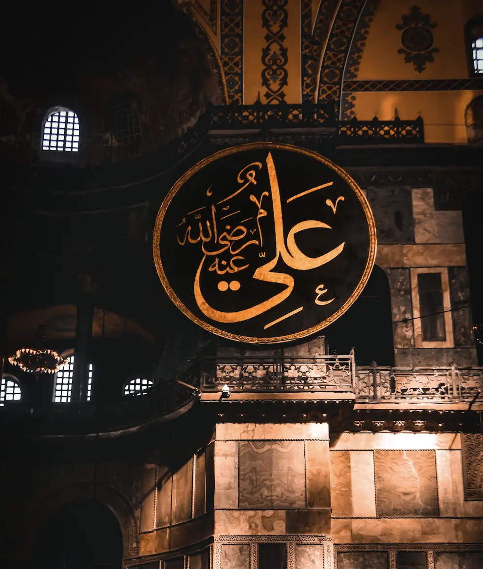 A large circular calligraphic medallion in gold script on a dark ground, framed in gilt and suspended in a domed interior above a stone gallery, giving the name Ali with the invocation radiya Allahu anhu