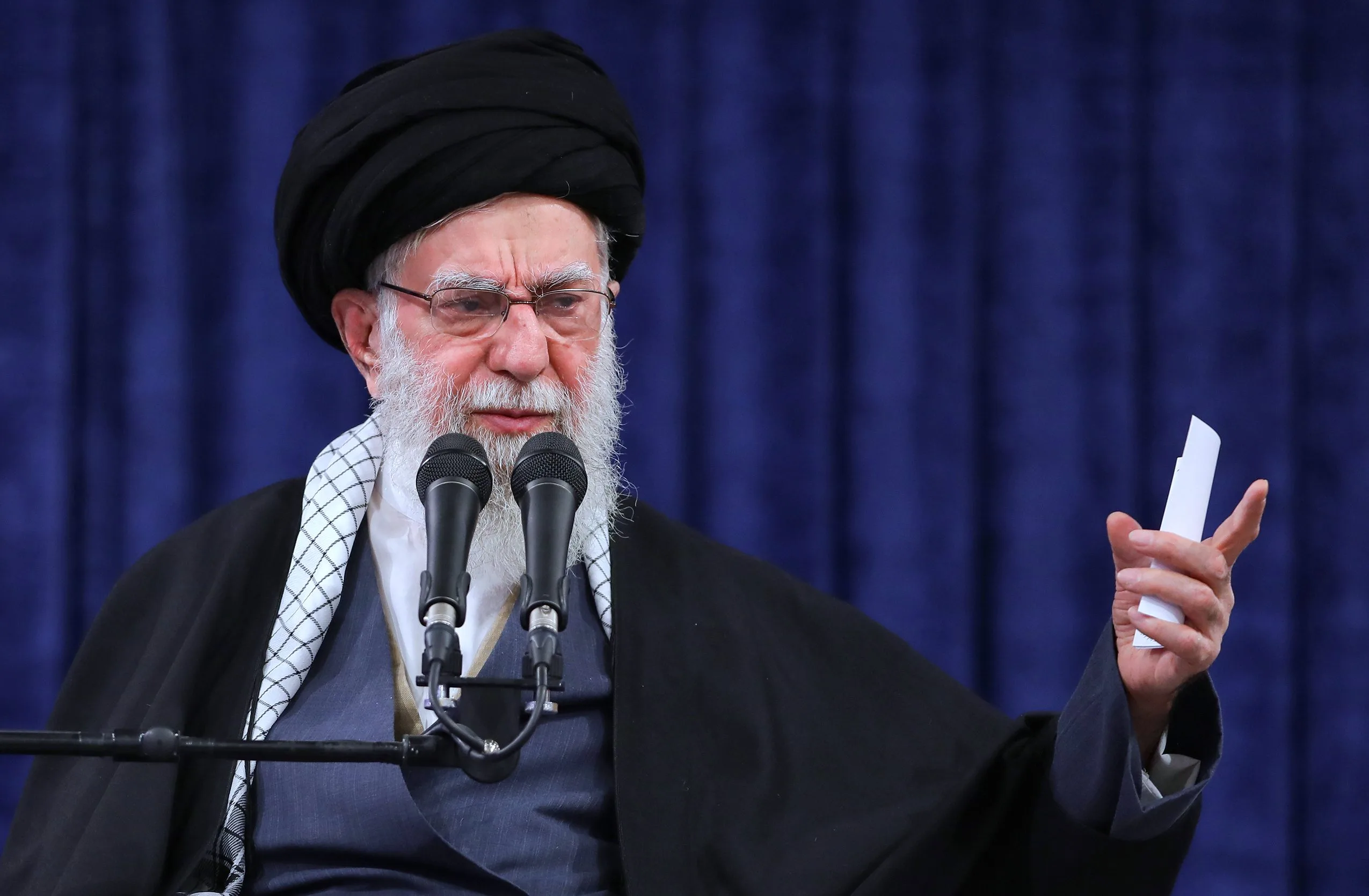 Photograph of an elderly bearded man in a black turban and dark cleric's robe with a checkered scarf and glasses, speaking at a pair of microphones while holding notes, against a blue backdrop