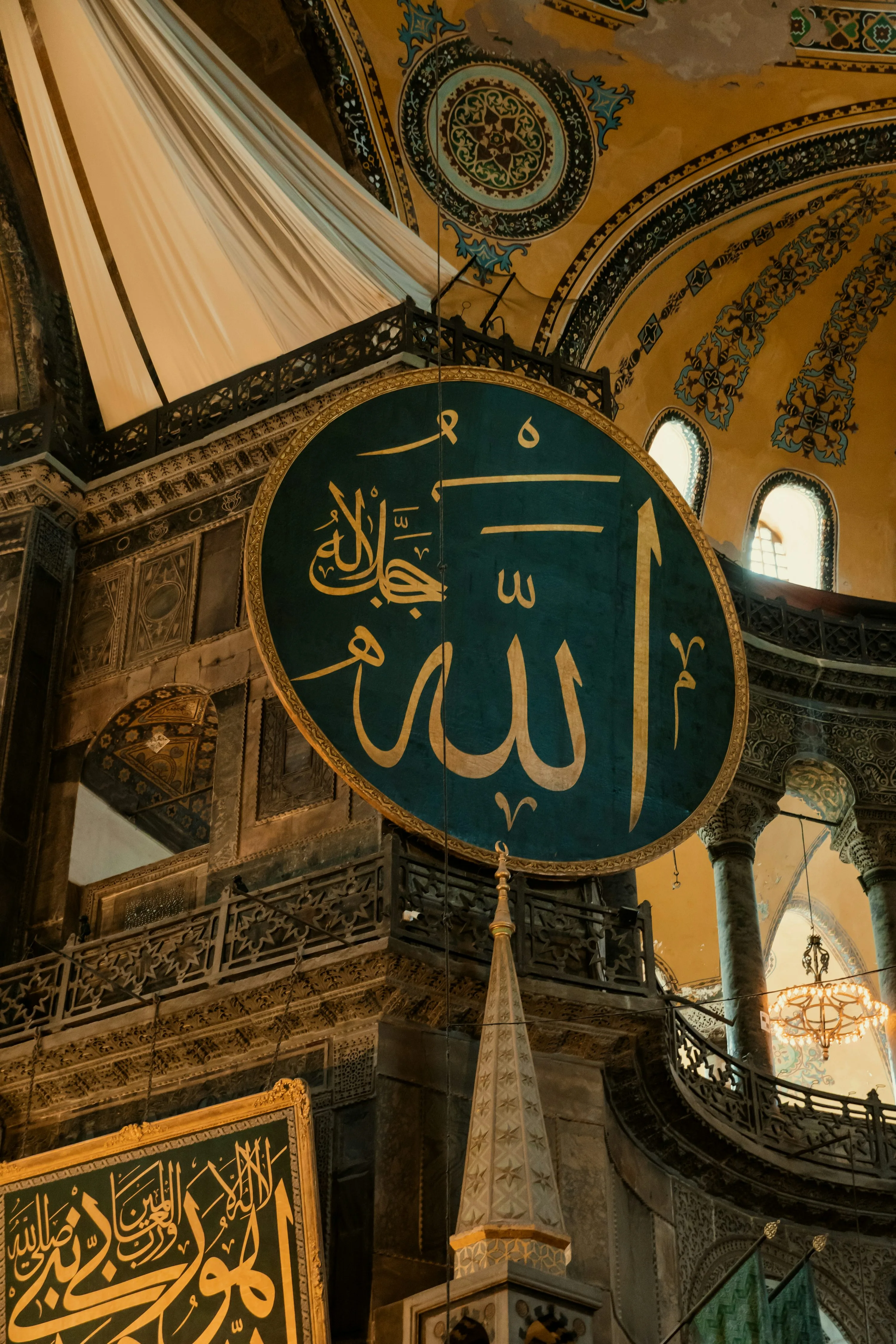 A large circular calligraphic medallion in gold script on a dark green ground, framed in gilt and suspended in the domed interior of Hagia Sophia above an ironwork gallery, bearing the name of Allah with a smaller invocation at the upper left, with painted Ottoman vault decoration and arched windows behind