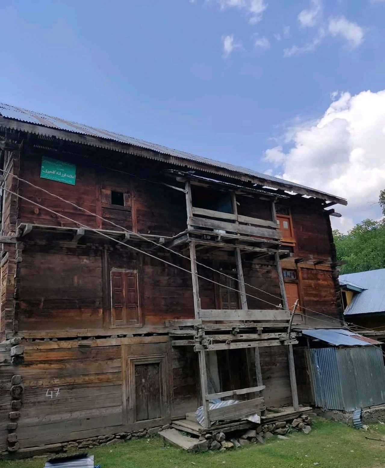 A tall three-storey house built of horizontally stacked weathered timber beams, with wooden shuttered windows, a projecting timber gallery frame and a corrugated metal roof, a green signboard lettered in Urdu fixed near the eaves at the left, and grass in the foreground