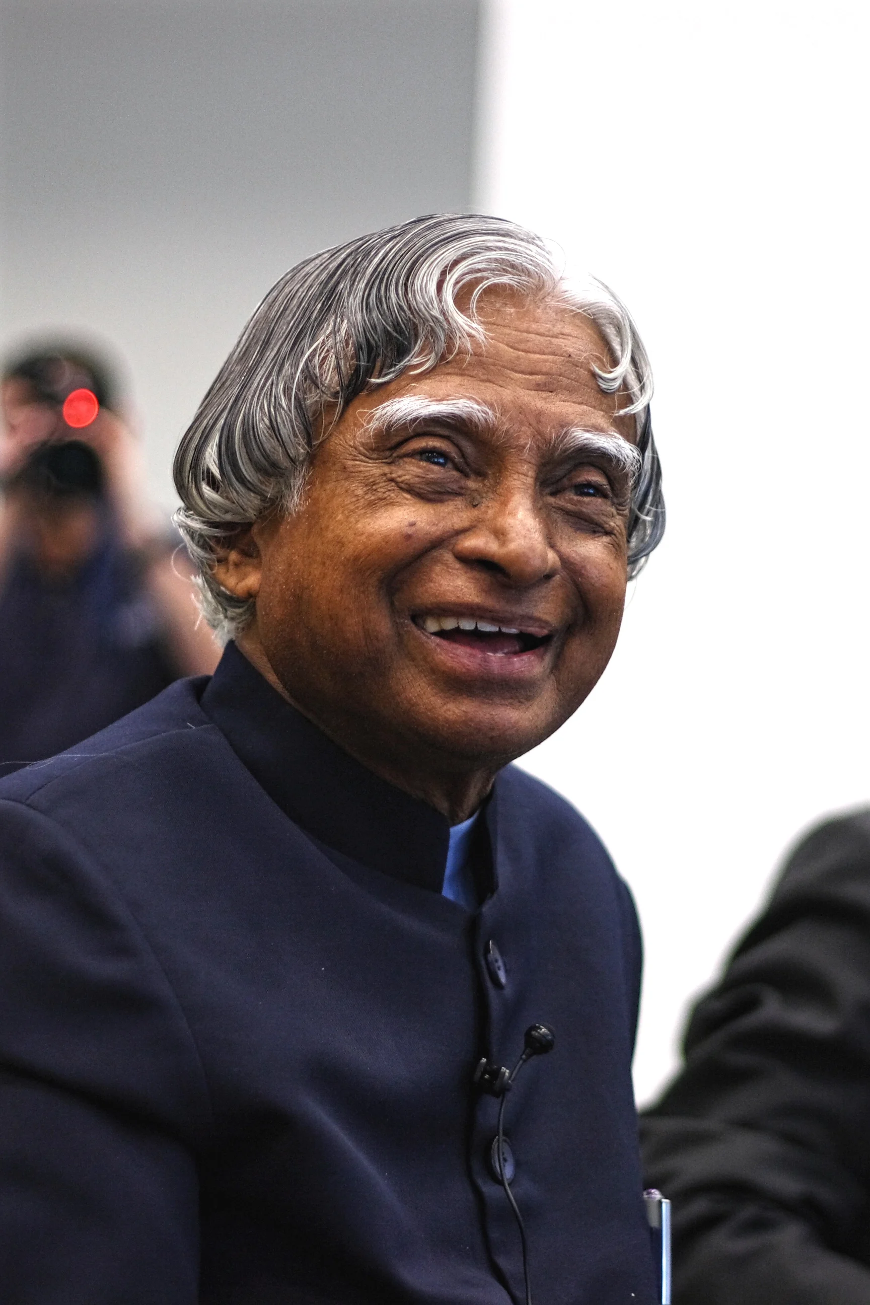 Colour photograph of an elderly man smiling broadly, with long silver-grey hair swept back from his forehead, wearing a dark navy closed-collar jacket over a pale blue shirt with a clip-on microphone at the chest; a photographer with a camera is out of focus behind him