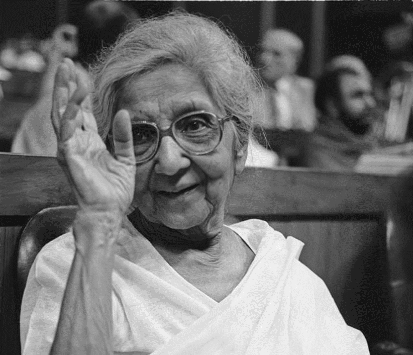 Black-and-white photograph of Aruna Asaf Ali in later life, wearing round spectacles and a white sari, raising a hand in greeting at a public gathering