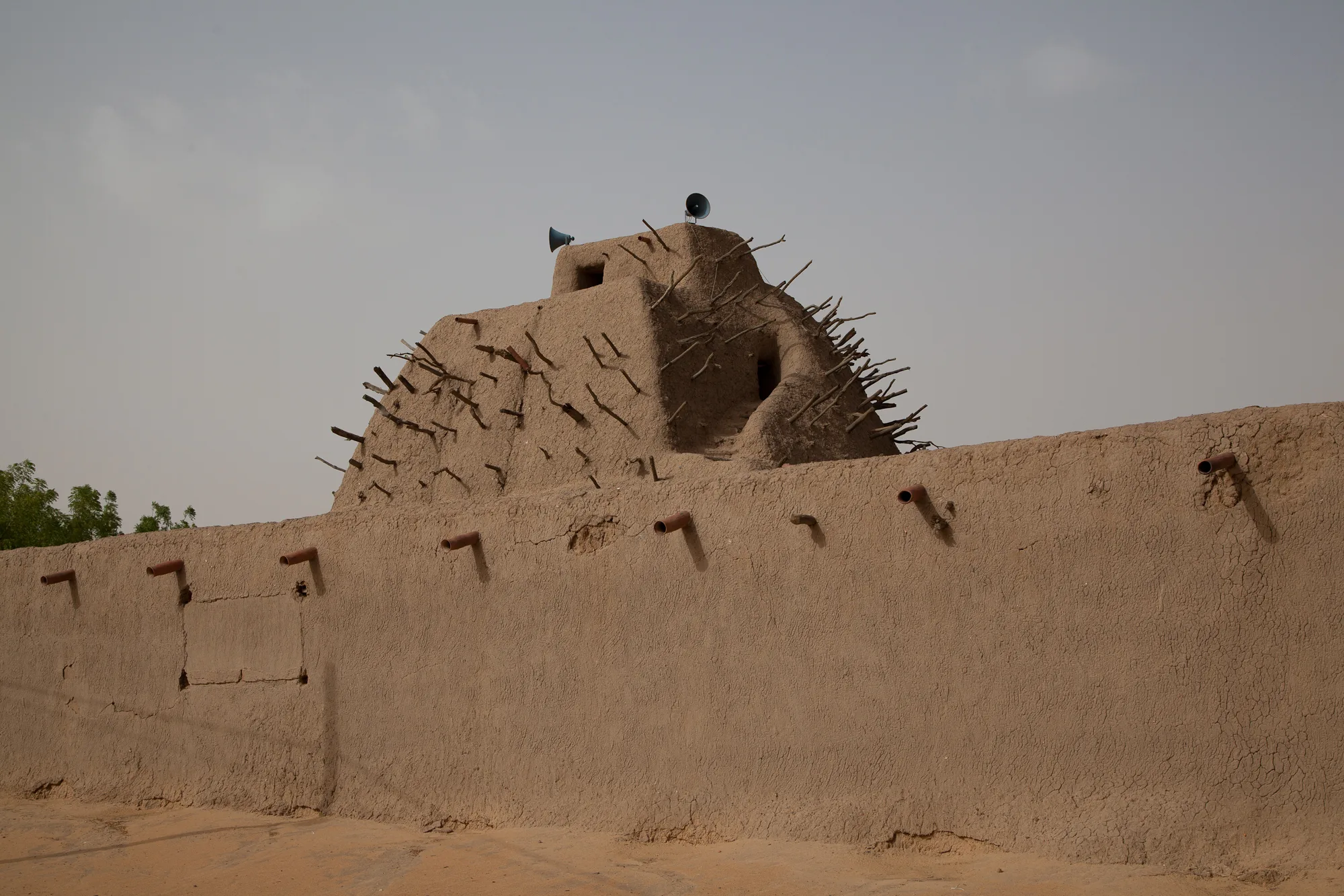 A tall stepped pyramid of mud brick, studded with projecting wooden beams and topped by two loudspeakers, rising behind a long mud-plastered wall set with clay drainpipes, under a grey sky
