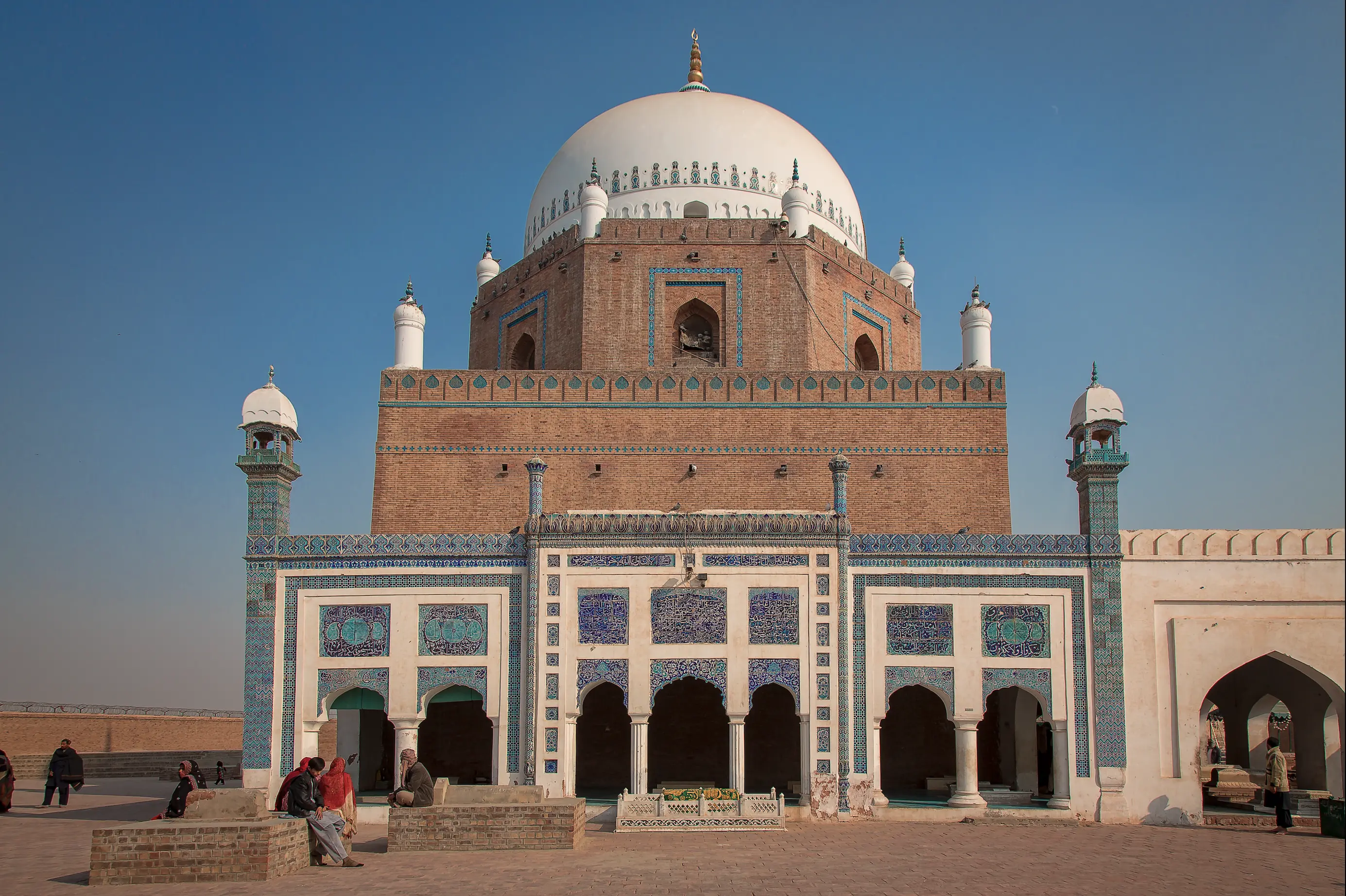 The shrine of Bahauddin Zakariya seen from its forecourt: a white hemispherical dome with a gilded finial above a square brick drum, set on tapering tiers of brickwork faced with turquoise and blue glazed tile, with four small white-domed corner turrets, and below it an arcaded screen carrying blue and white calligraphic tile panels; a few visitors cross the paved court