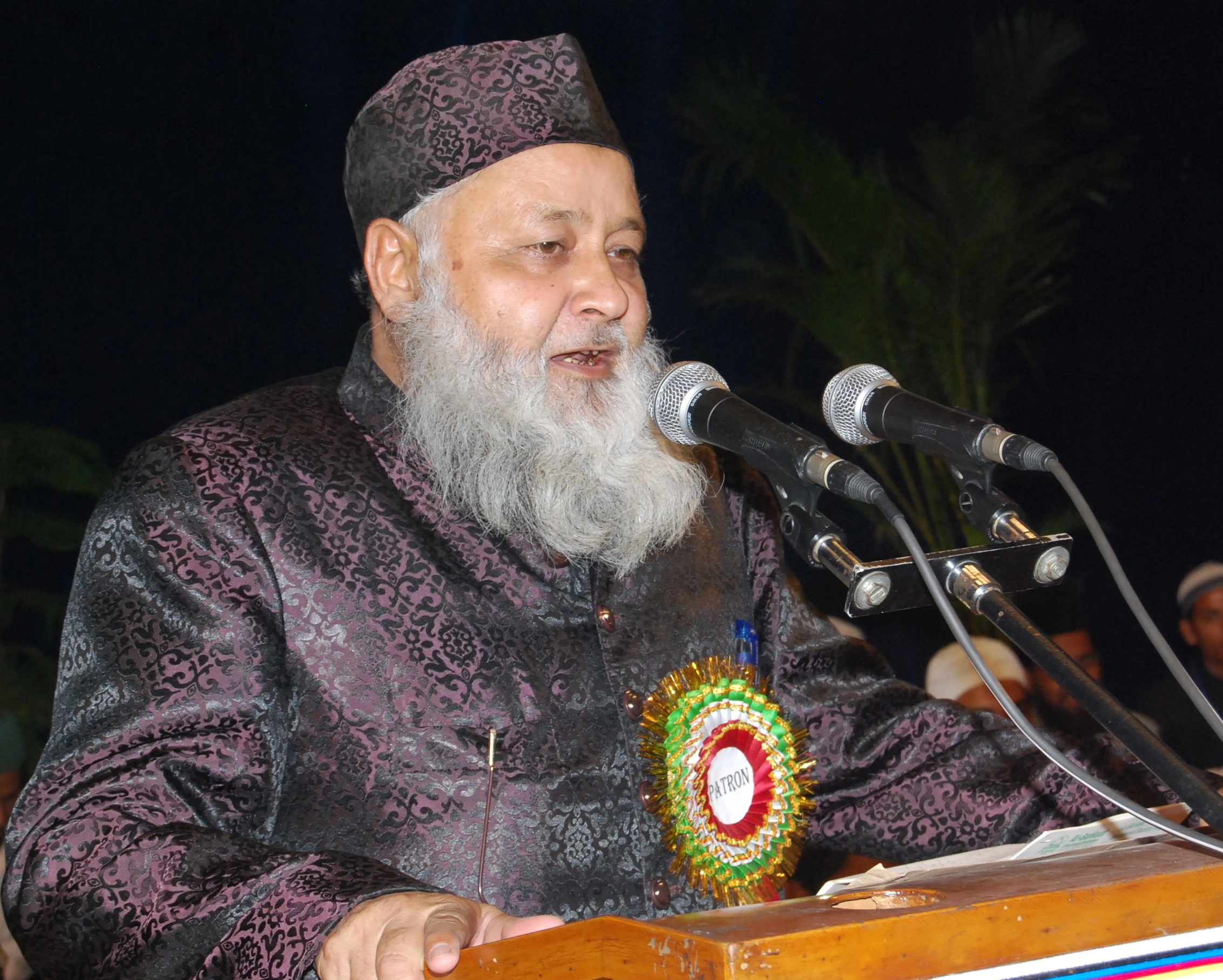 An elderly scholar with a white beard, wearing an embroidered cap and a patterned brocade sherwani with a rosette badge pinned to it, speaking at a wooden lectern fitted with two microphones at an evening gathering