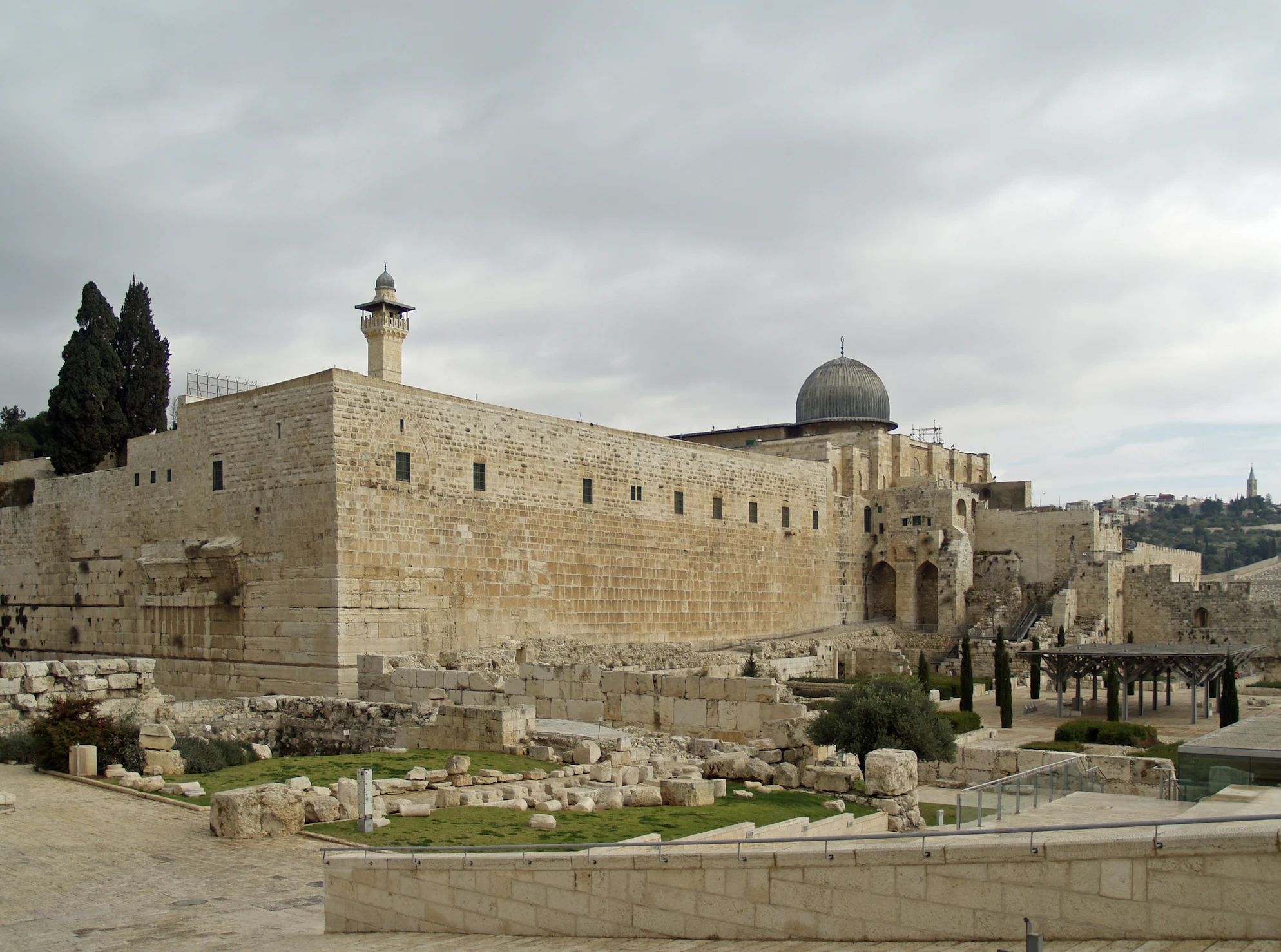 The ancient stone southern wall of the Haram al-Sharif in Jerusalem, with a minaret and the grey-lead dome of the Al-Aqsa Mosque rising above, and archaeological ruins in the foreground