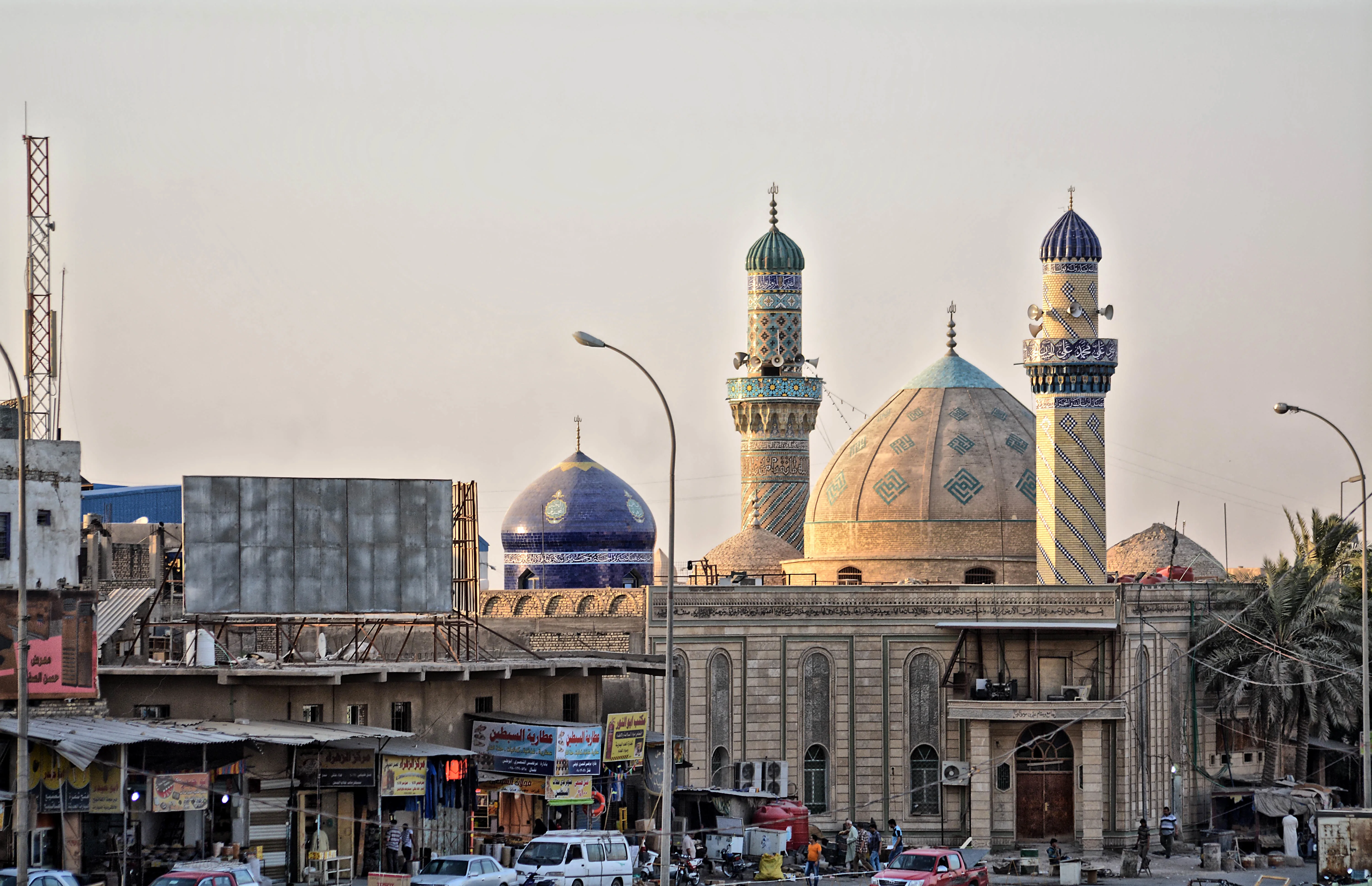 Tiled domes and two patterned minarets of a mosque in Basra rising above a street of shopfronts, signboards and parked vehicles