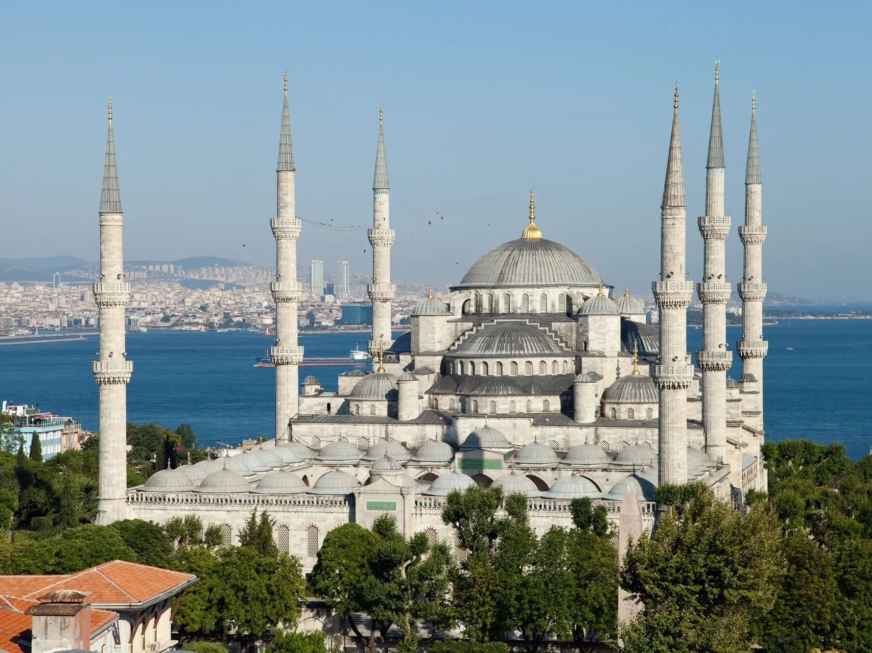 The Sultan Ahmed Mosque seen from higher ground: a central lead-grey dome with a gilded finial above a cascade of semi-domes and smaller domes, ringed by six slender fluted minarets with balconies, trees in the foreground and the sea and the far shore of Istanbul behind