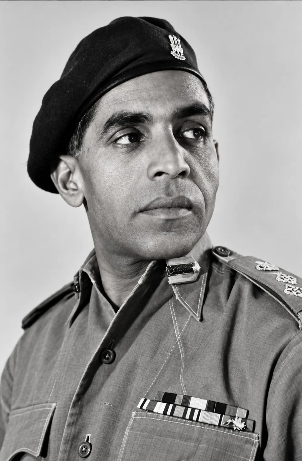 Black-and-white portrait photograph of a clean-shaven man in army uniform looking to one side, wearing a dark beret with a metal cap badge, an open-collared shirt with a gorget patch at the throat, rank insignia on the shoulder strap, and a row of medal ribbons above the left breast pocket