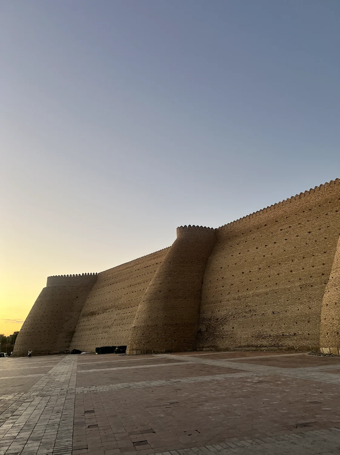 The massive sloping brick walls and crenellated ramparts of the Ark citadel of Bukhara at sunset, with a wide brick-paved plaza in front