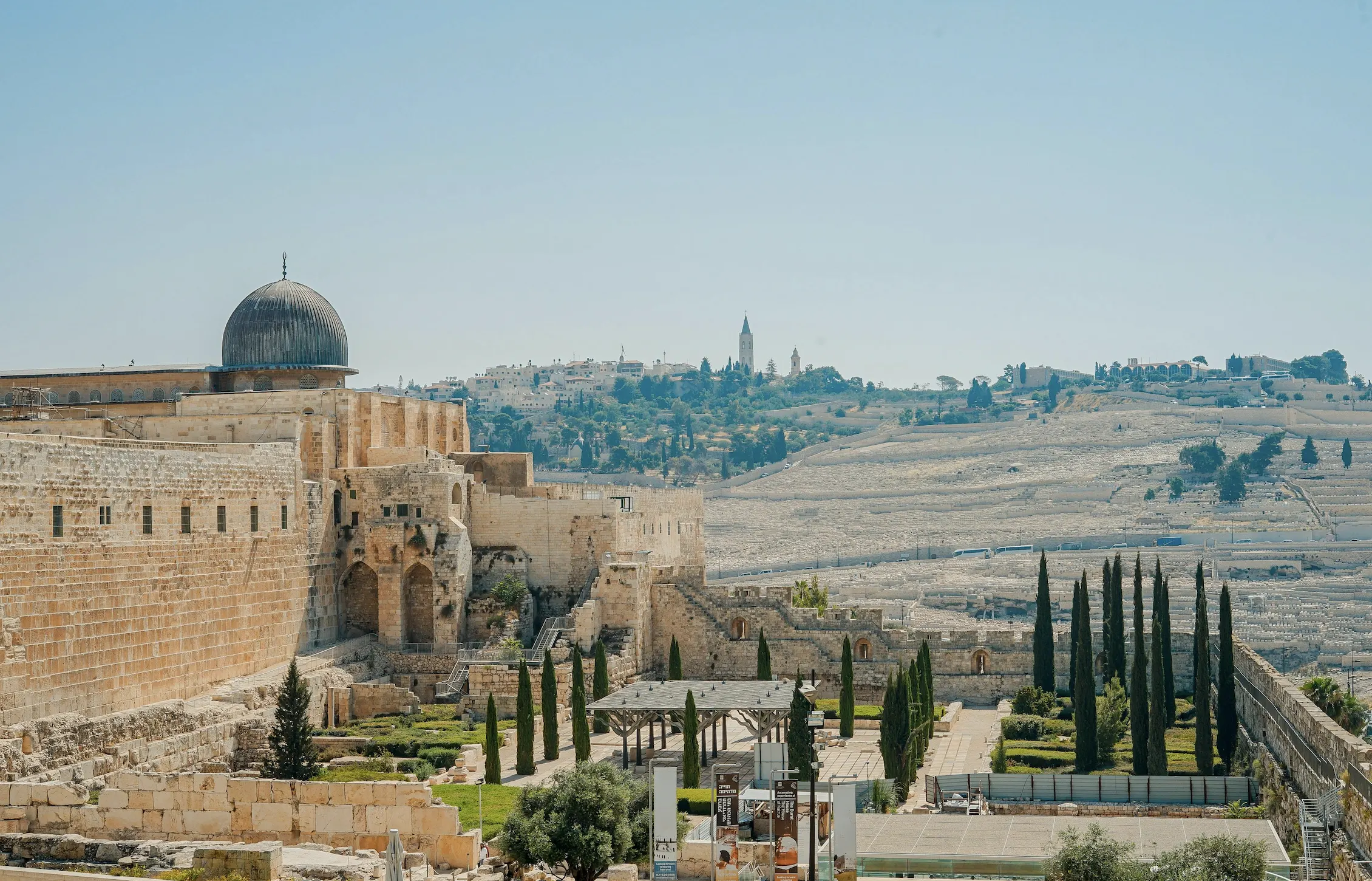 A wide view of the Old City of Jerusalem showing the silver-domed Al-Aqsa Mosque above the ancient stone walls of the Temple Mount, with the Mount of Olives and cypress trees beyond