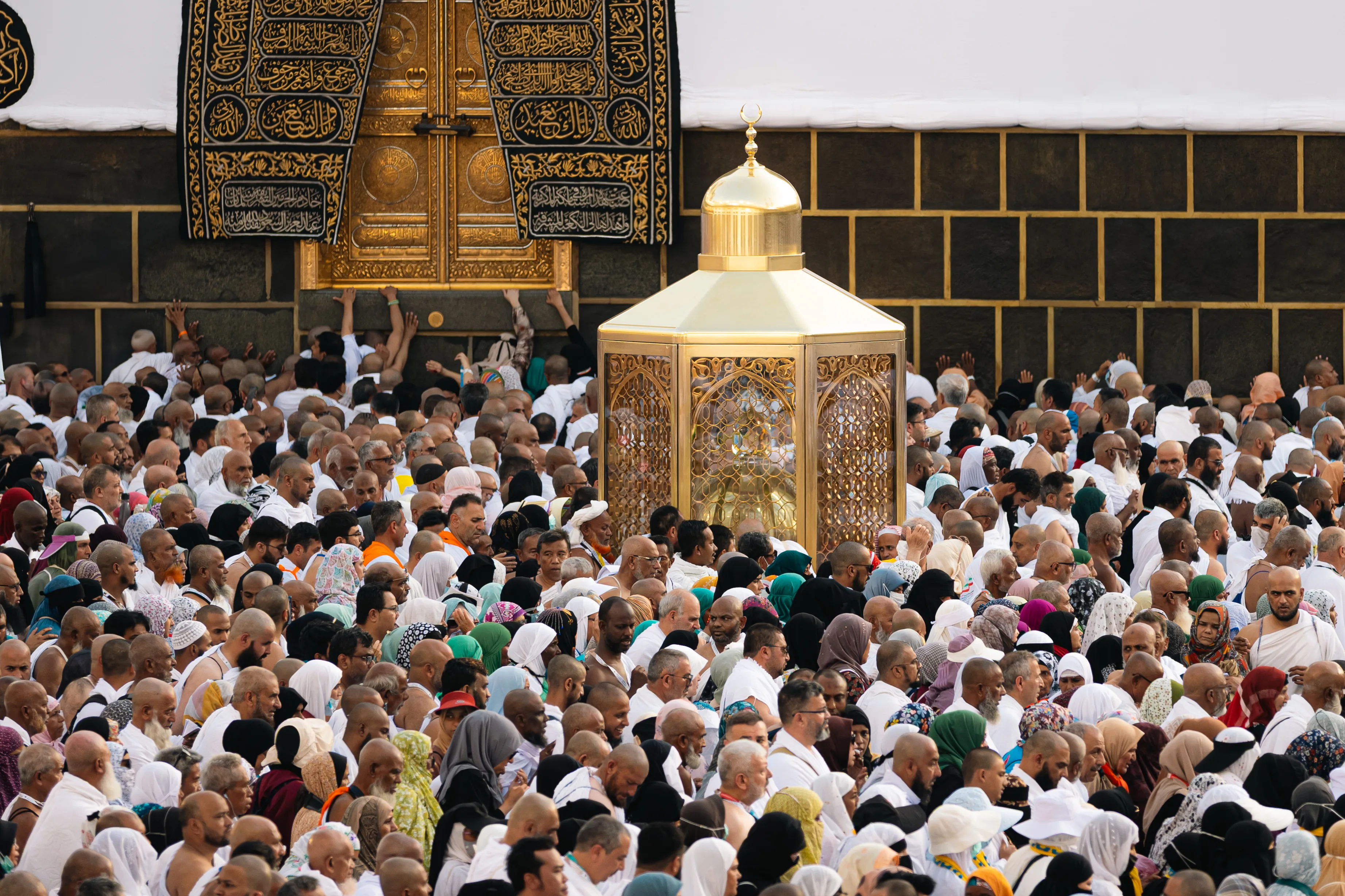 The gilded door of the Kaaba set in its black kiswah embroidered with gold Qur'anic calligraphy, with the golden Maqam Ibrahim enclosure and a crowd of pilgrims before it