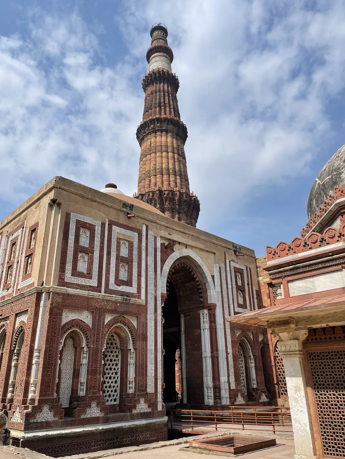 The Qutb Minar rising behind the carved red-sandstone and white-marble Alai Darwaza gateway in the Qutb complex, Delhi, under a partly cloudy sky