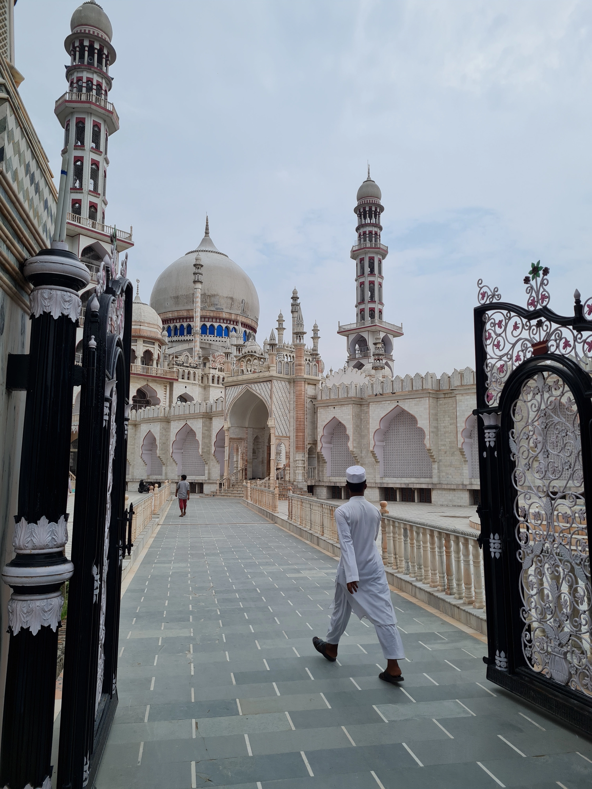 A white marble mosque and seminary complex with a large central dome and tall minarets banded in red, seen through a pair of open wrought-iron gates, with a student in a white kurta and cap walking along the paved approach