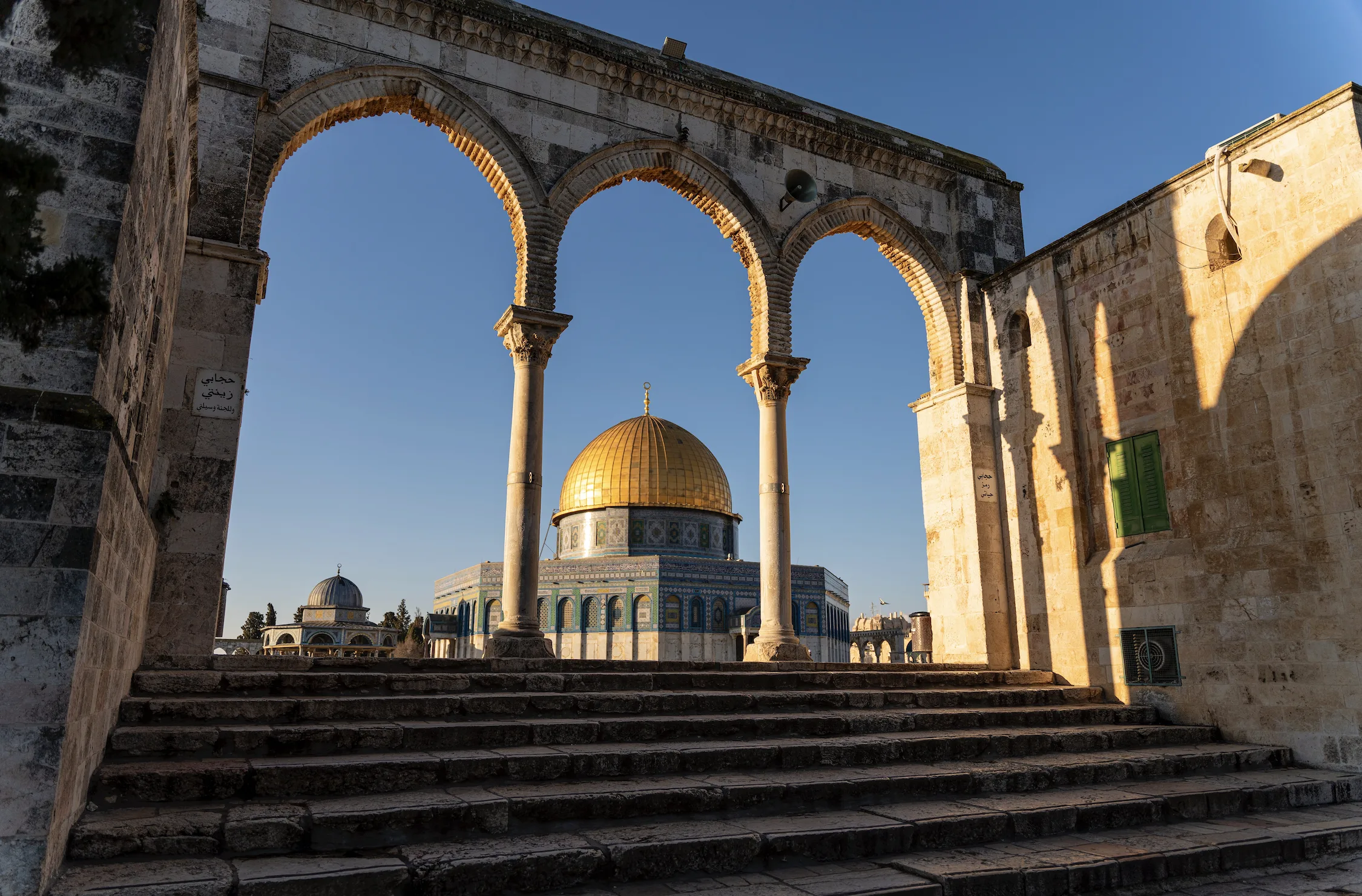 The Dome of the Rock in Jerusalem seen through a stone arcade, its golden dome rising above the blue-tiled octagonal drum, with steps leading up to the platform