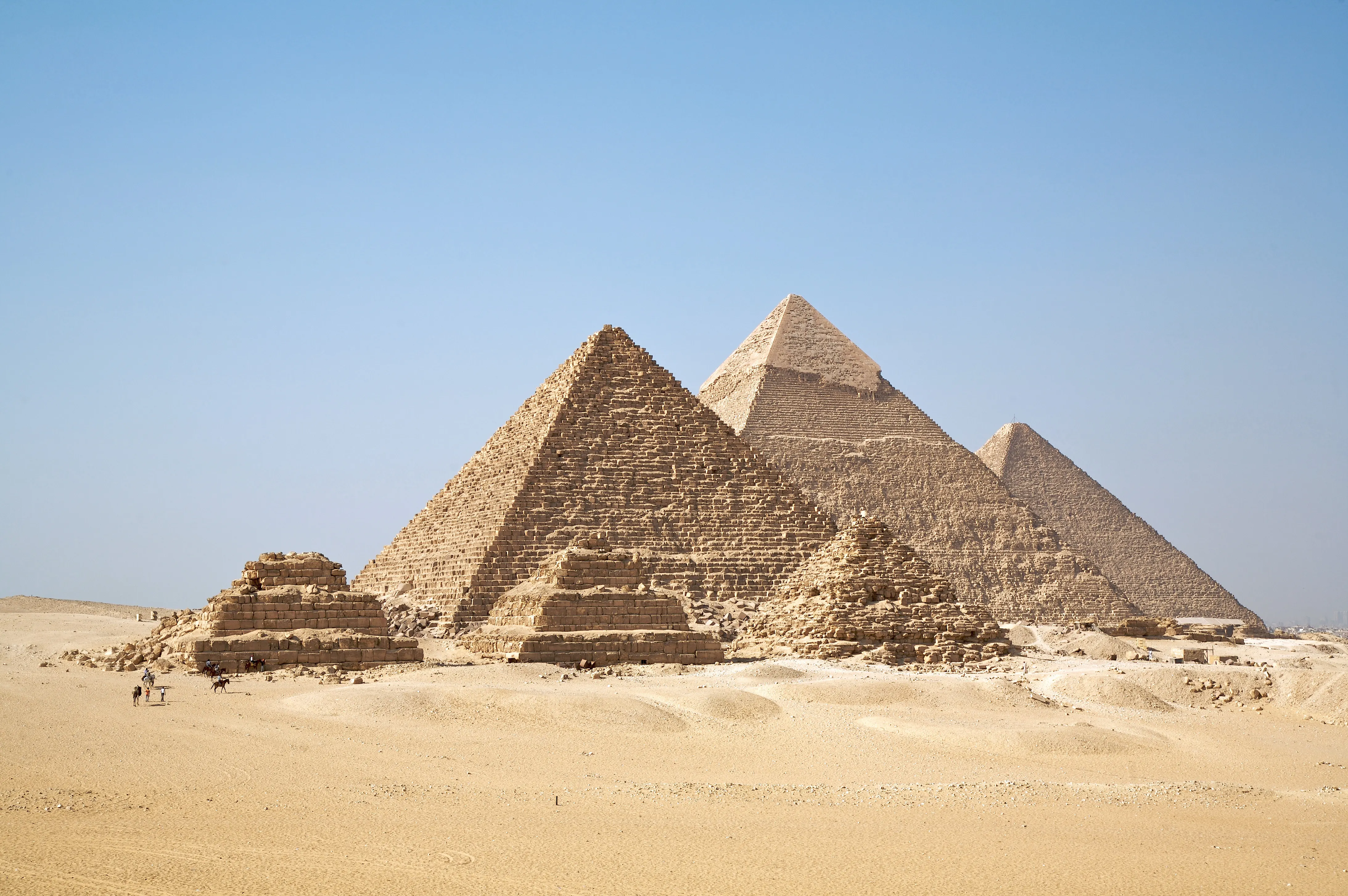 The three pyramids of Giza rising from the desert plateau under a clear sky, with smaller ruined structures in the foreground