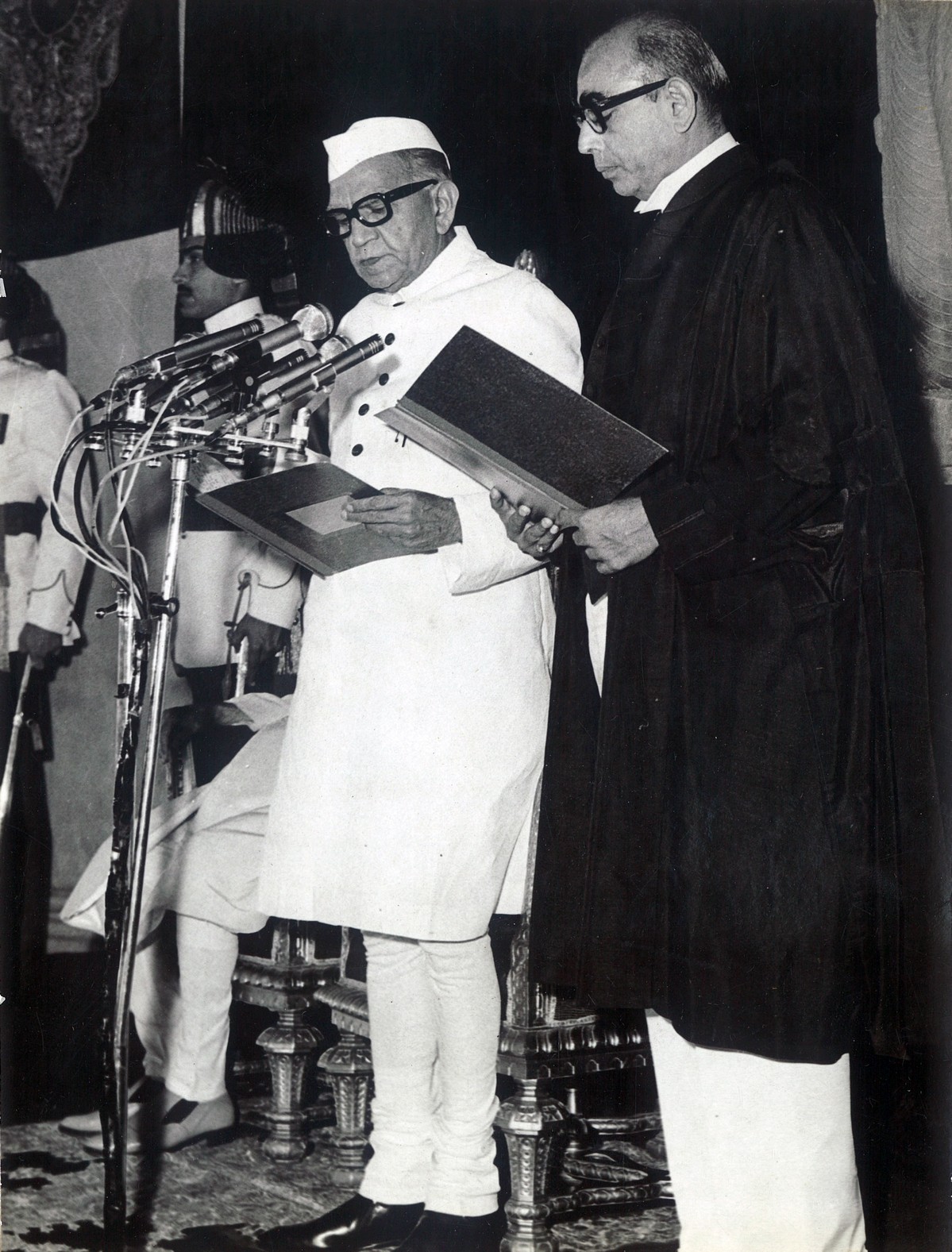 Black-and-white photograph of Fakhruddin Ali Ahmed, on the left in a white achkan and white cap, reading the oath at a bank of microphones, with a man in dark judicial robes standing beside him and ceremonial guards behind