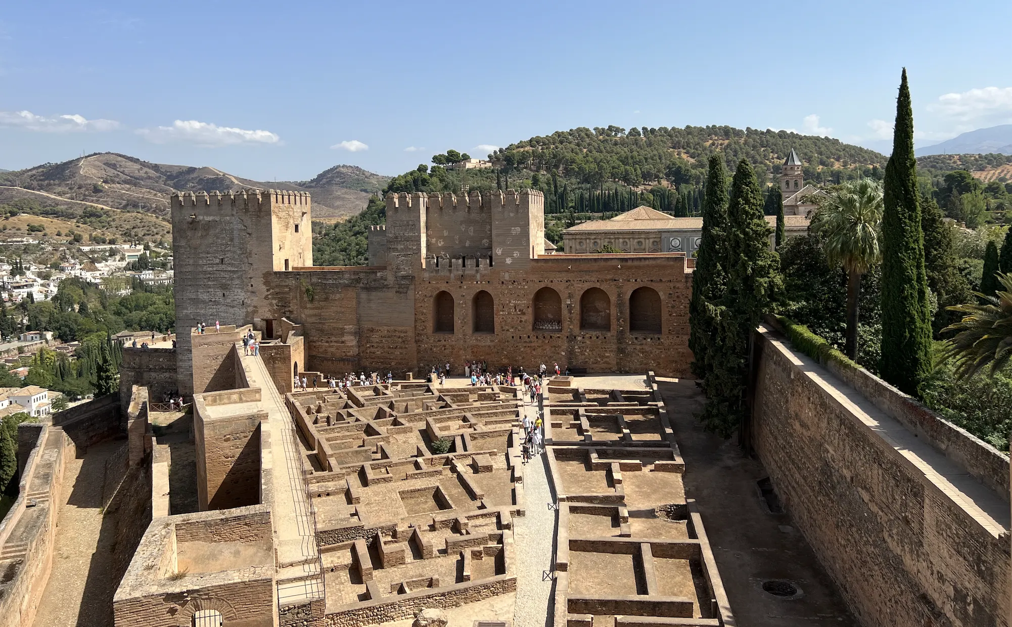 The Alcazaba fortress of the Alhambra, with crenellated stone towers and ramparts above the excavated foundations of the old military quarter, hills and the city of Granada beyond