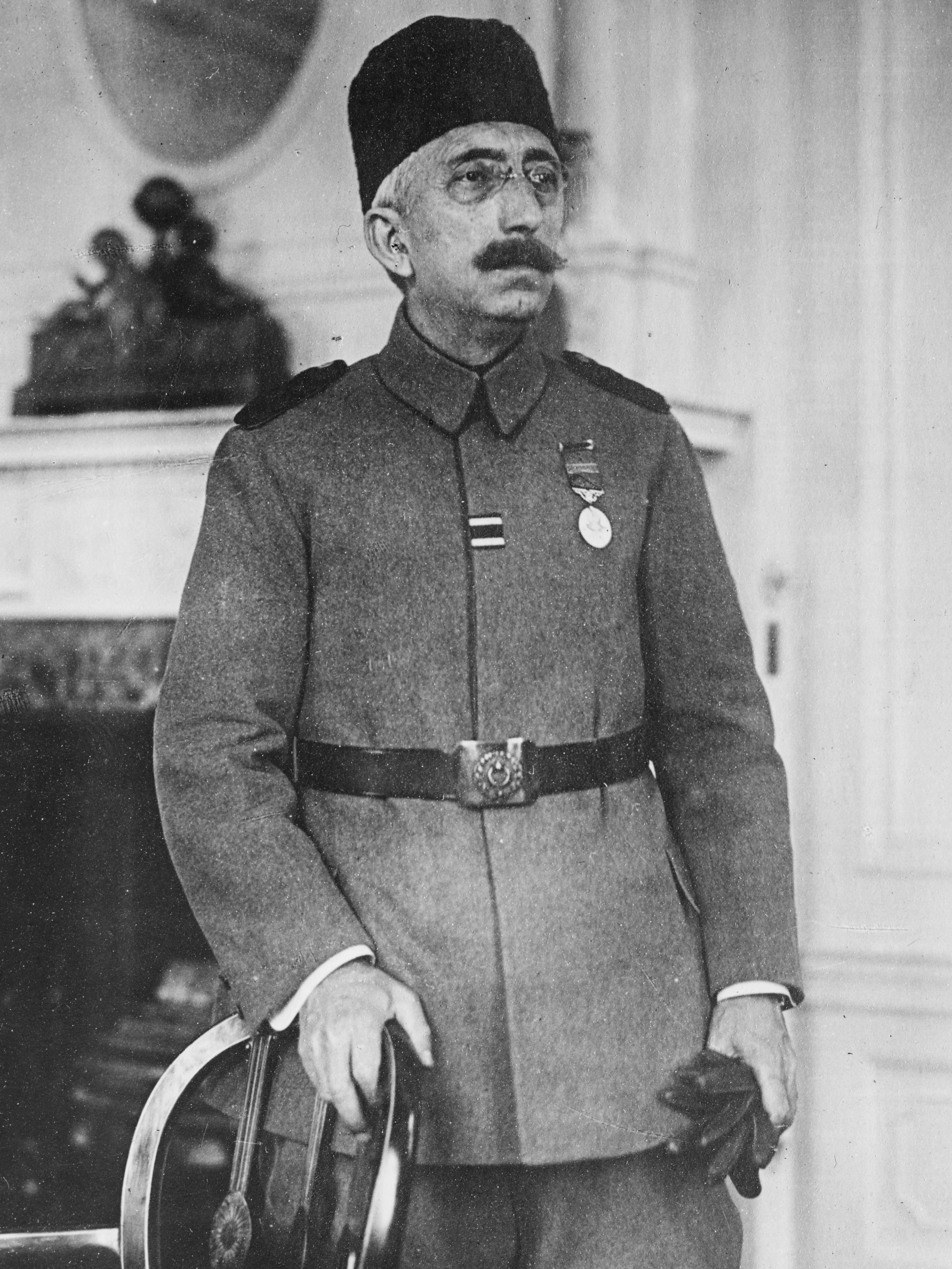 A black and white photograph of Mehmed VI in military uniform, wearing a dark kalpak and spectacles, a medal at his chest and a sword at his side