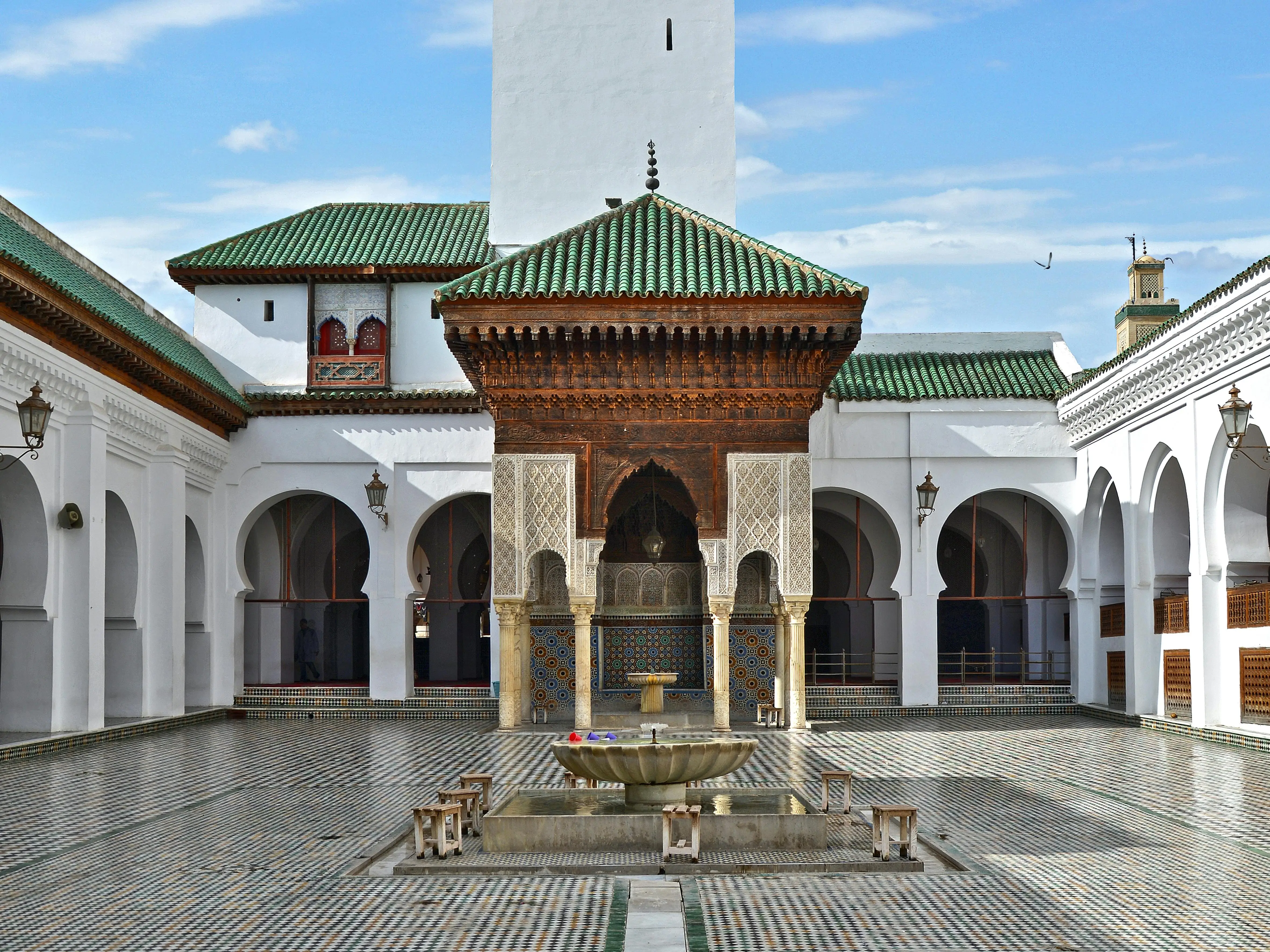 Courtyard of the al-Qarawiyyin mosque and university in Fez, with whitewashed horseshoe-arch arcades, green-tiled roofs, a carved wooden fountain pavilion, and a central marble fountain on a patterned tiled floor