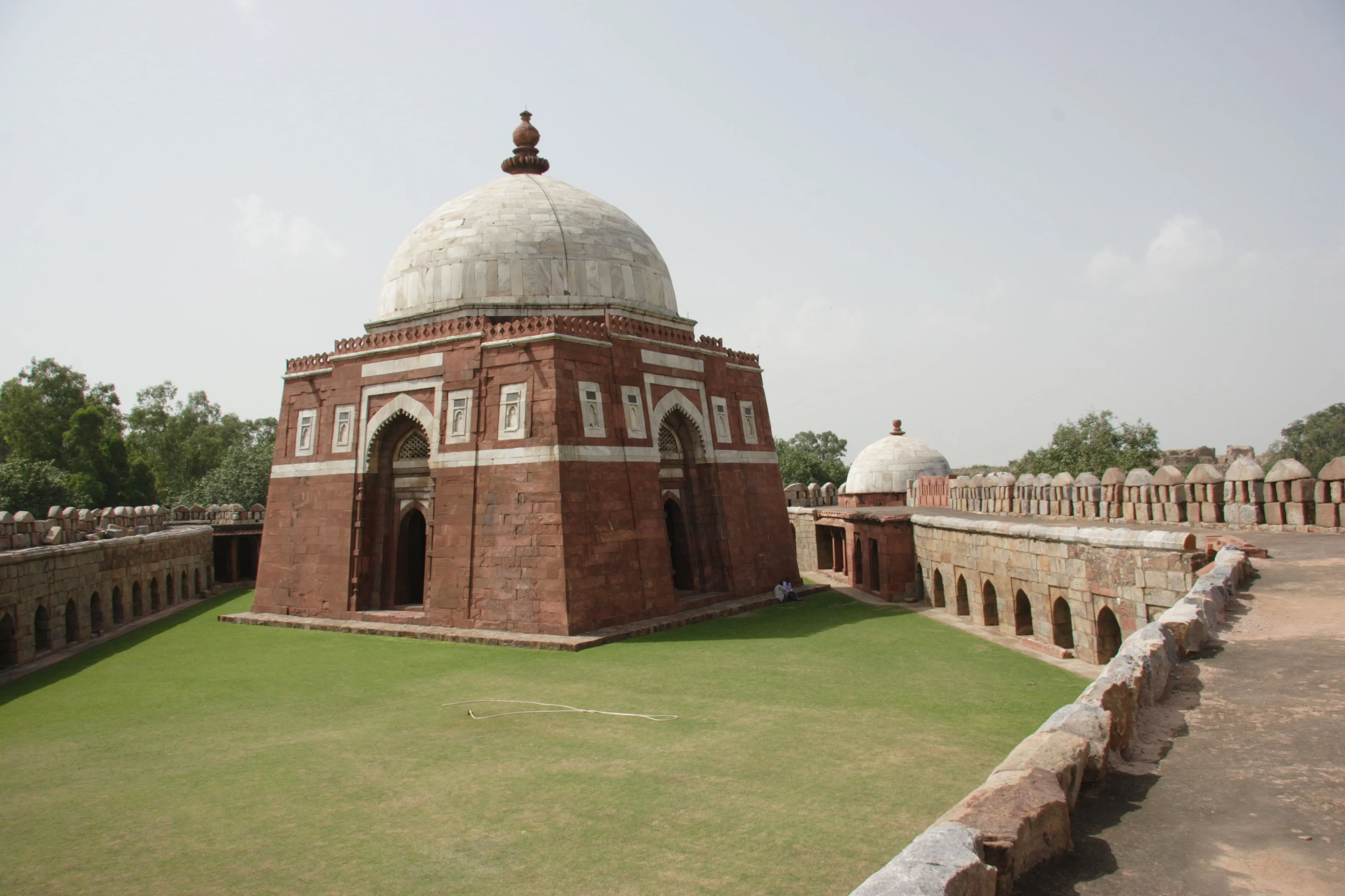 The mausoleum of Ghiyath al-Din Tughluq, a red sandstone tomb banded with white marble and crowned by a white dome, standing within a walled enclosure of arcades and battlements