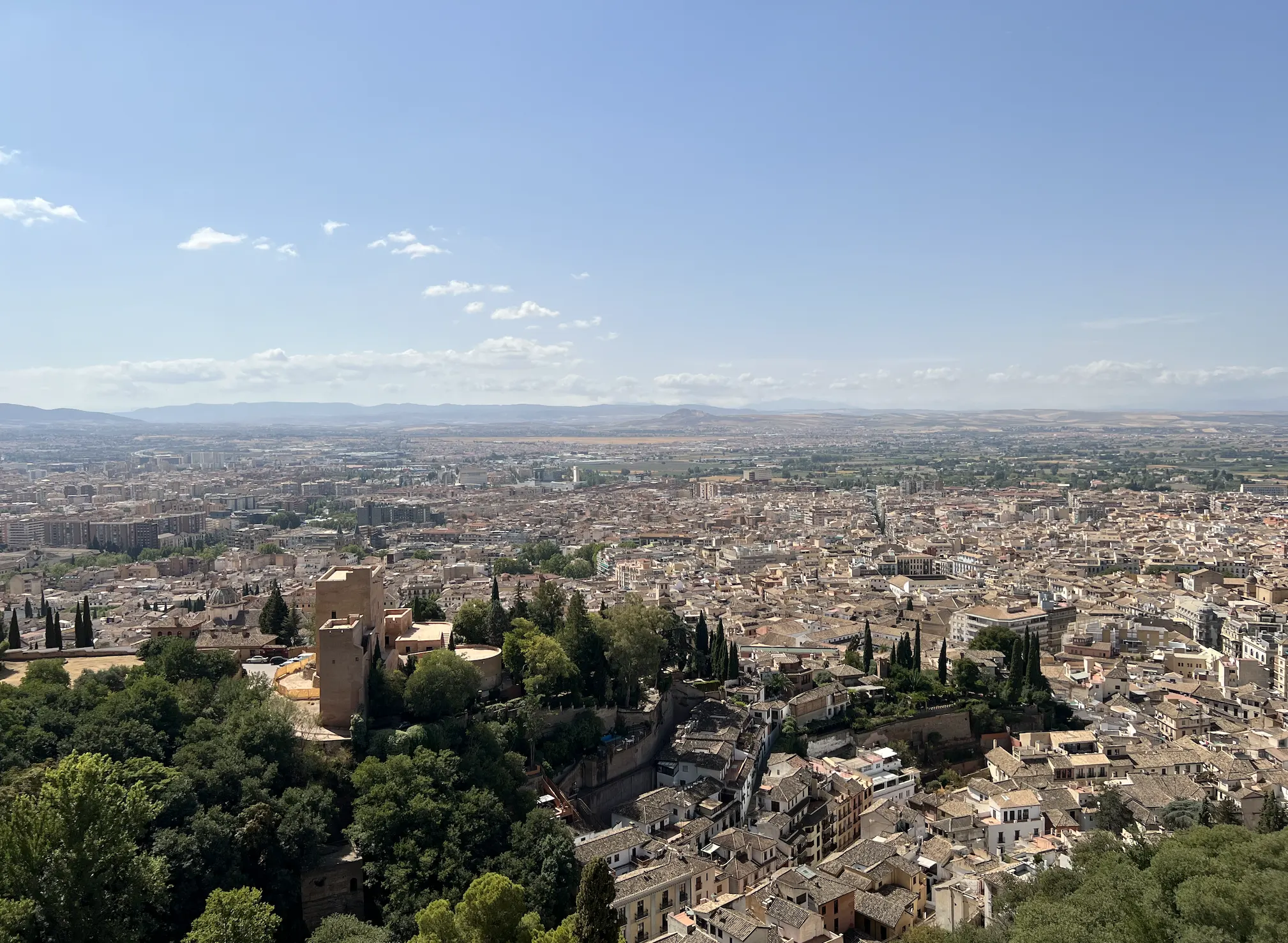 Panoramic view over the city of Granada from above, red-tiled rooftops stretching to the plain and distant mountains, with a stone tower and cypress trees in the foreground