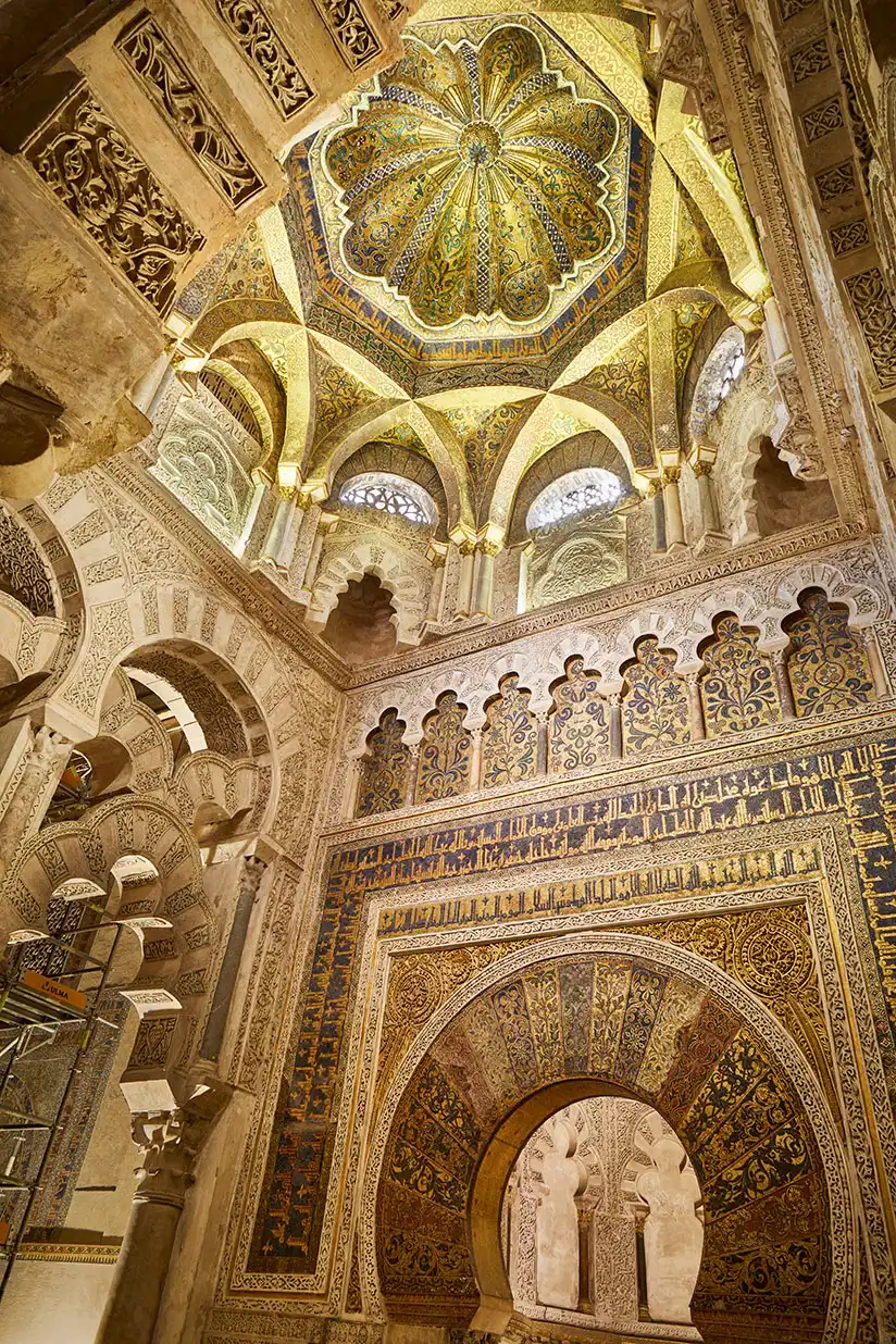 The mihrab of the Great Mosque of Cordoba beneath a ribbed golden mosaic dome, the horseshoe-arch niche framed by gold-ground Arabic inscription and intricate stucco