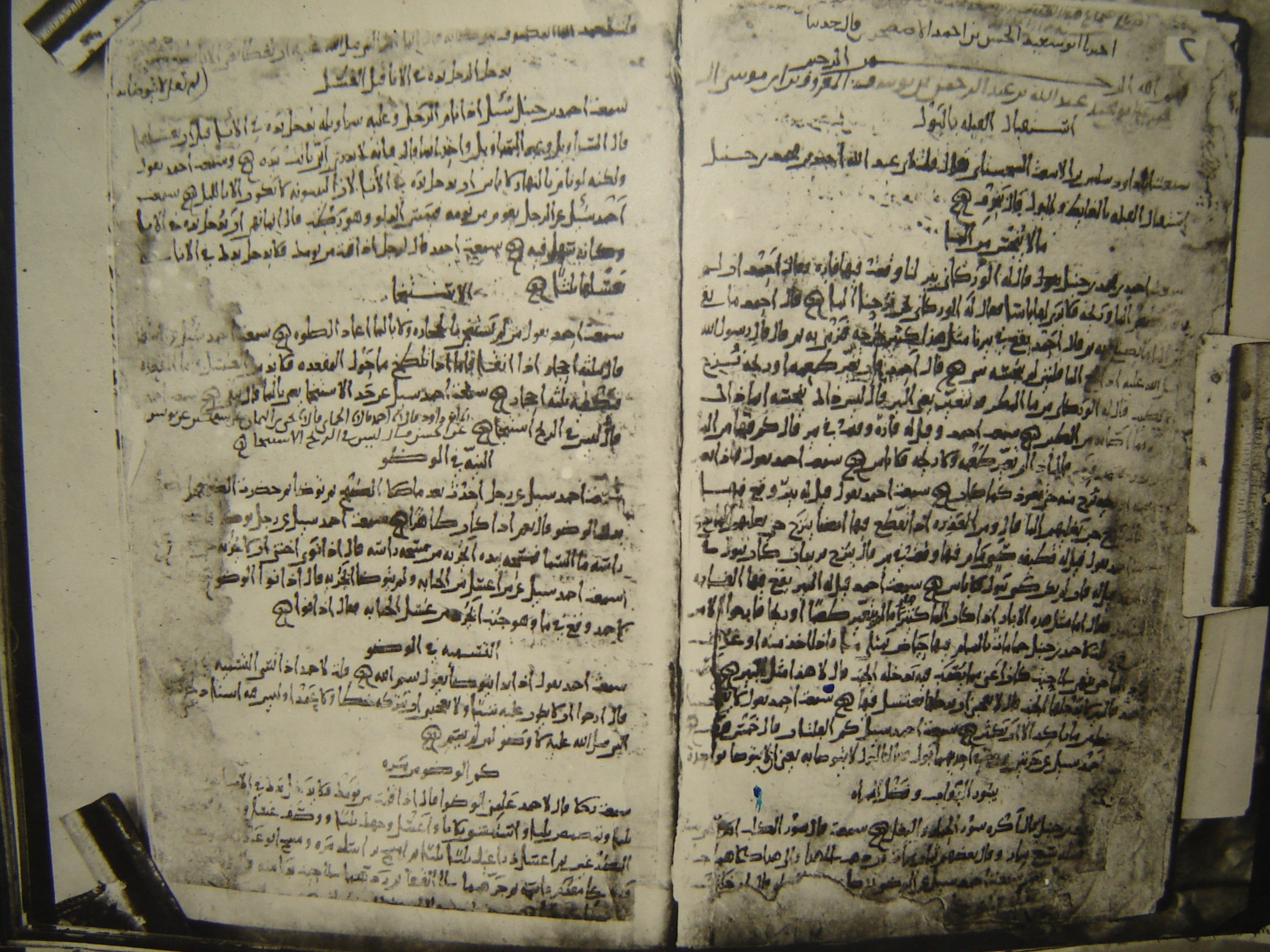 A photograph of two facing pages of an early Arabic manuscript, worn and stained, closely written in dark ink with chapter headings and marginal additions, held open by clips