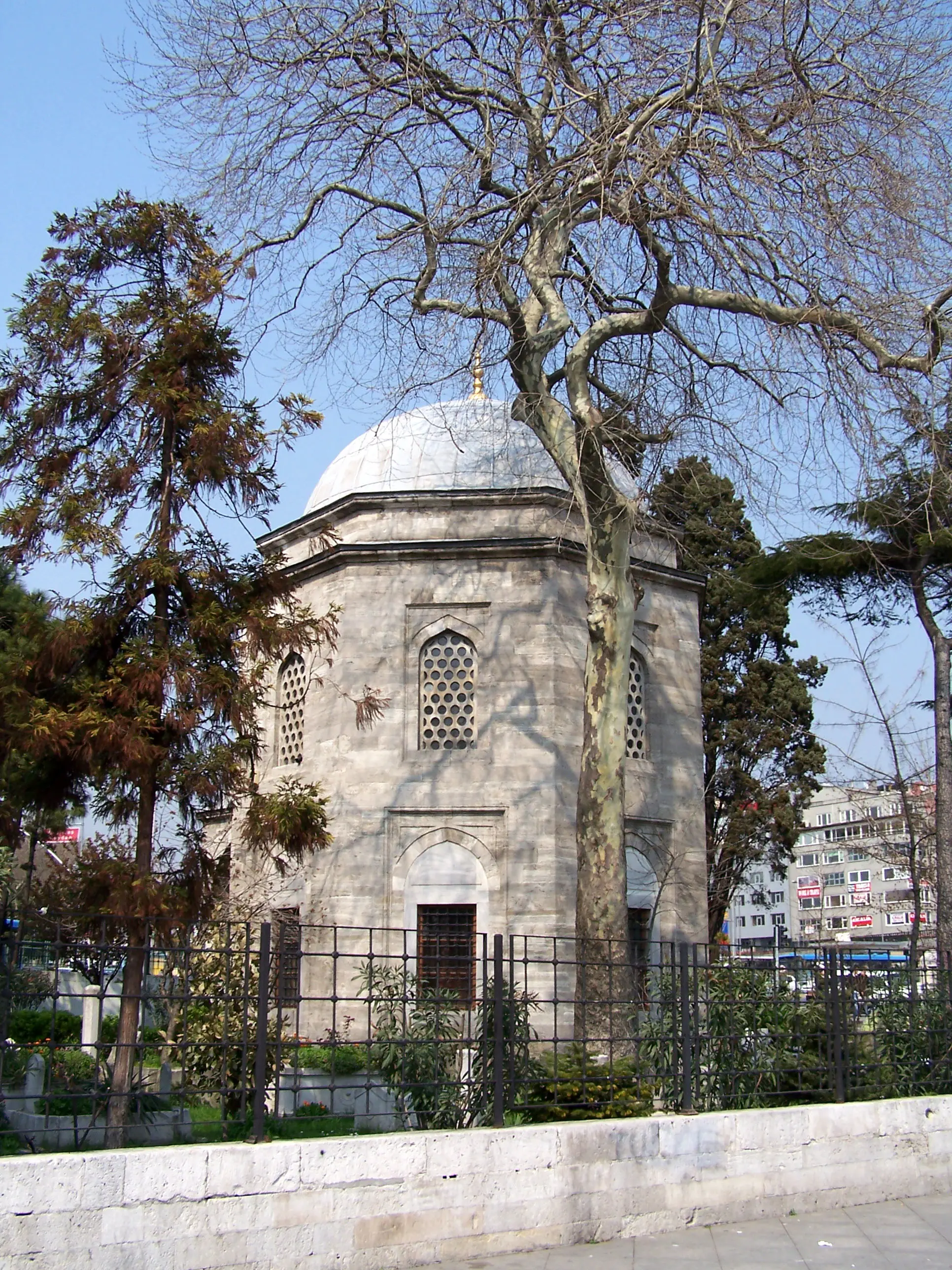 An octagonal stone tomb with a lead-covered dome and latticed windows, enclosed by an iron railing and shaded by trees