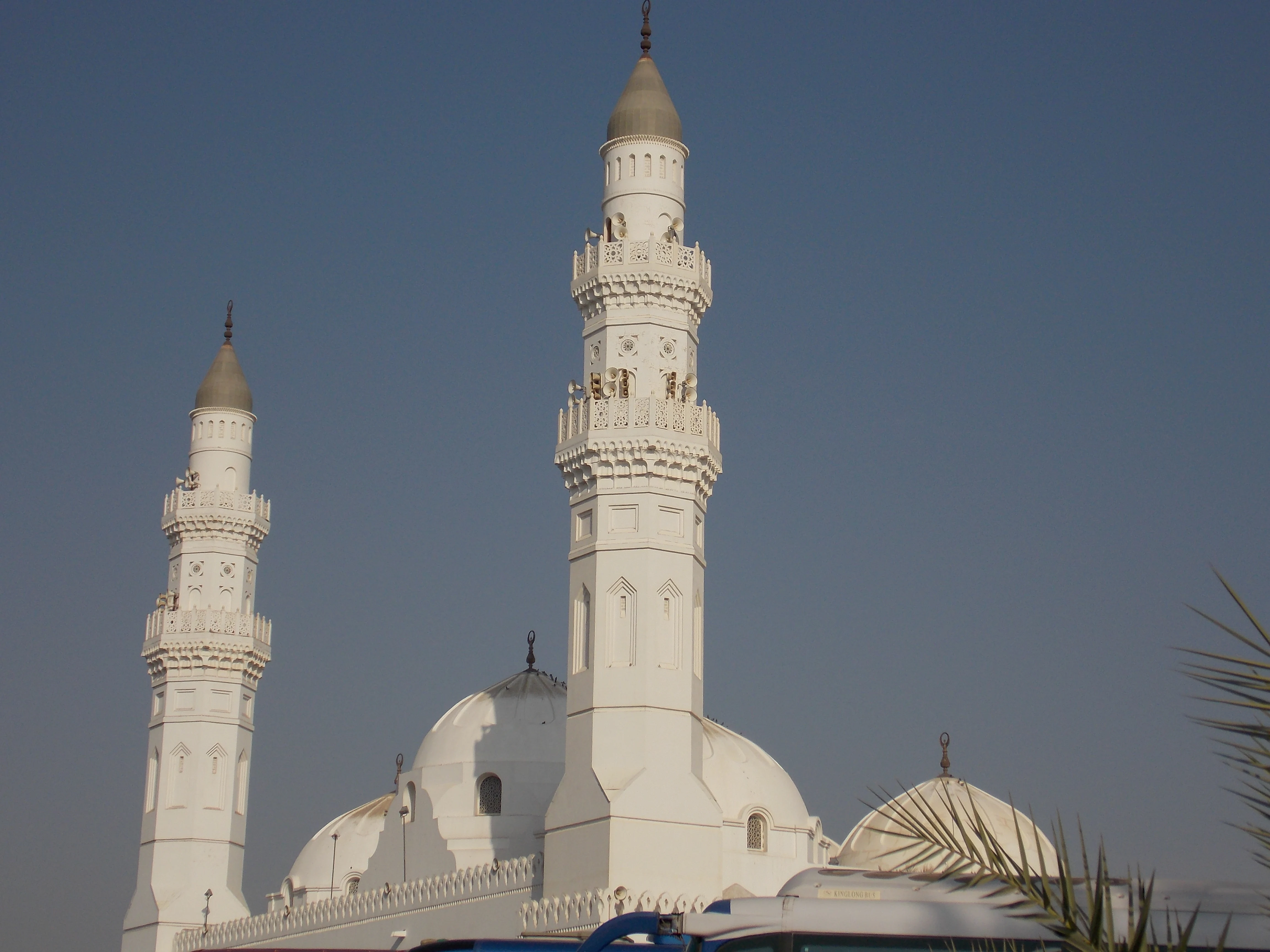 The white Quba Mosque in Medina, with two tall minarets and a cluster of white domes, seen over the roof of a bus with palm trees in front