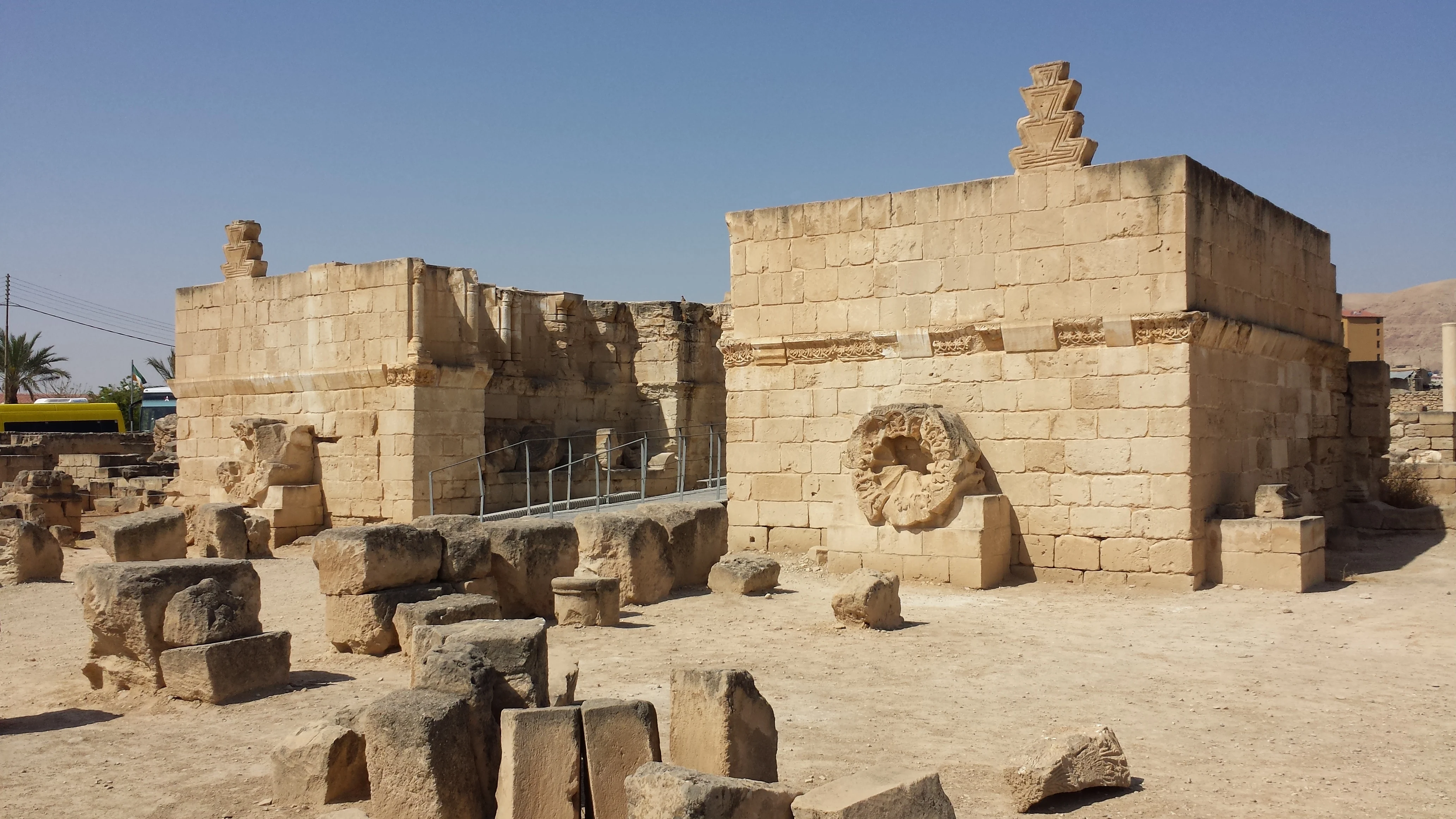 Ashlar limestone walls of a ruined palace standing to full height, crowned with stepped merlons and carved with a cornice frieze and a large stone rosette, with column drums and fallen blocks lying across the excavated ground in front