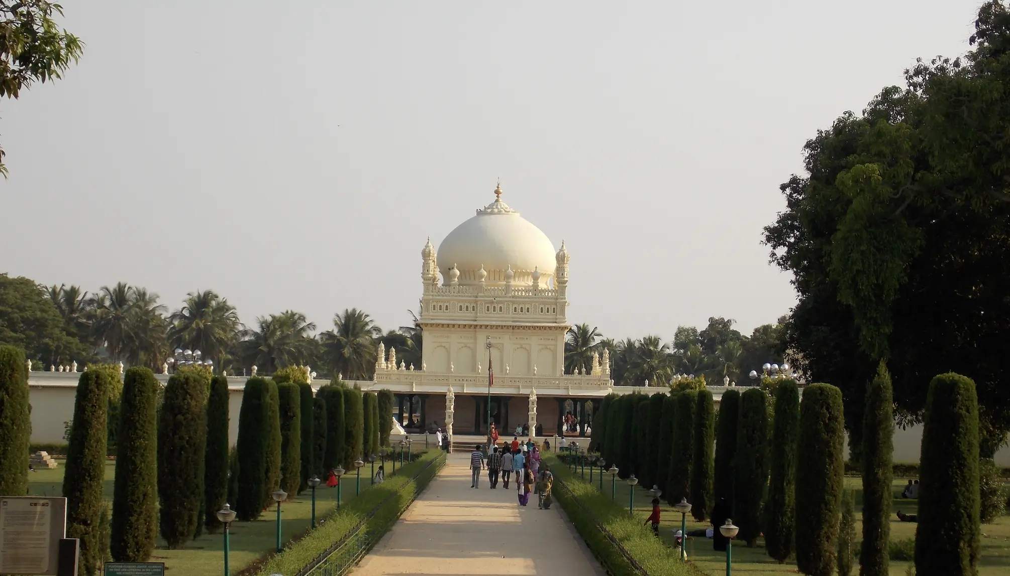 Colour photograph of the Gumbaz at Srirangapatna seen down a formal garden avenue lined with clipped conical trees, the white domed mausoleum with corner minarets rising above a colonnaded terrace, with visitors walking the path towards it