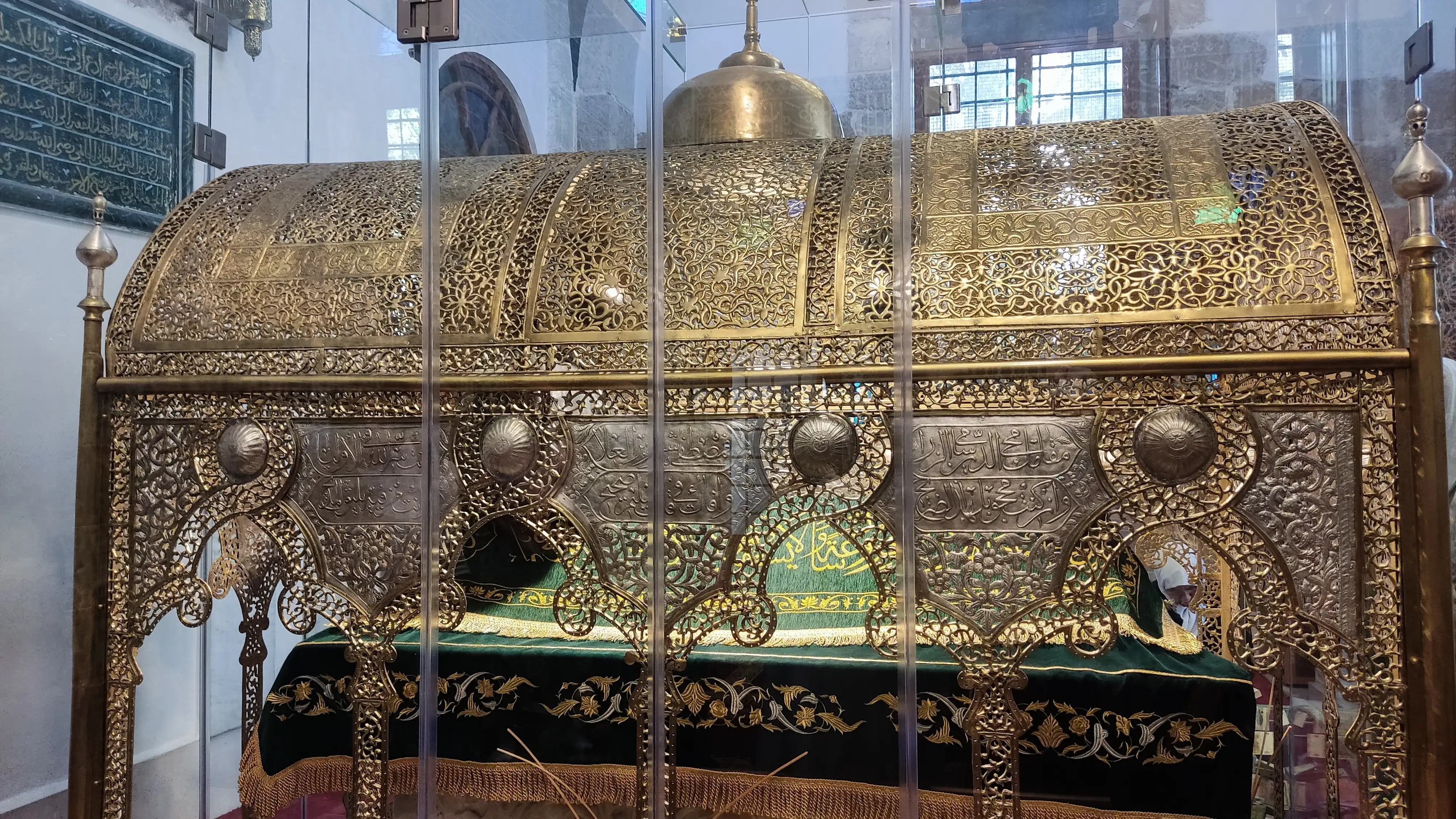 A cenotaph draped in green cloth embroidered in gold, enclosed by an ornate gilded screen of pierced metalwork behind glass, with a calligraphic panel on the wall at the upper left