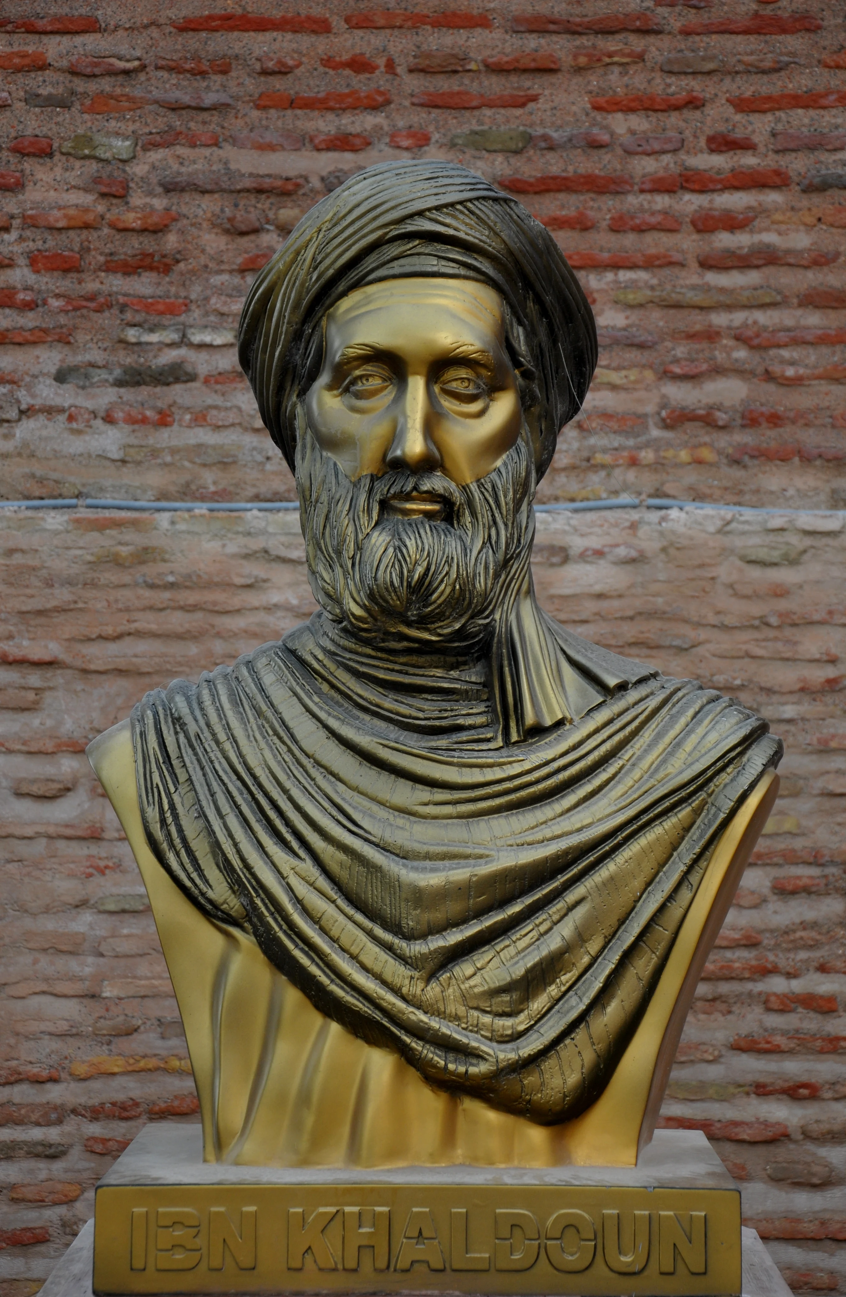 A gold-coloured bust of a bearded man in a turban, on a plinth inscribed IBN KHALDOUN, against a red brick wall