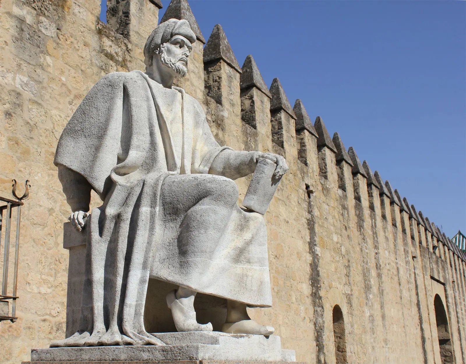 A stone statue of a seated, bearded man in a turban and long robes, holding a book, set on a plinth beside the crenellated stone walls of Córdoba