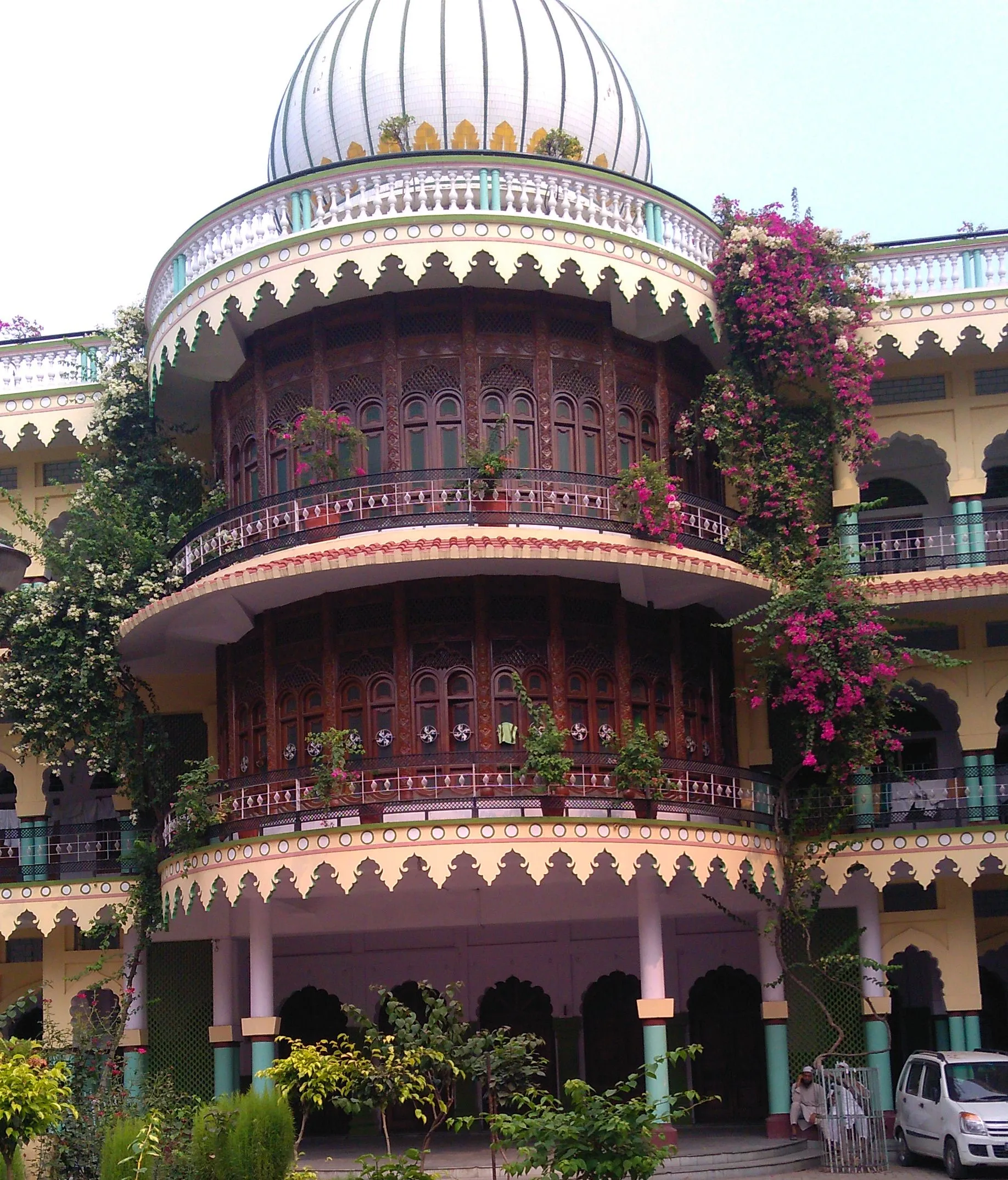 A semicircular projecting bay of carved dark wood with arched lattice windows rising through two storeys, crowned by a white ribbed dome on a balustraded drum, with scalloped arch decoration in cream and turquoise and bougainvillea climbing the facade