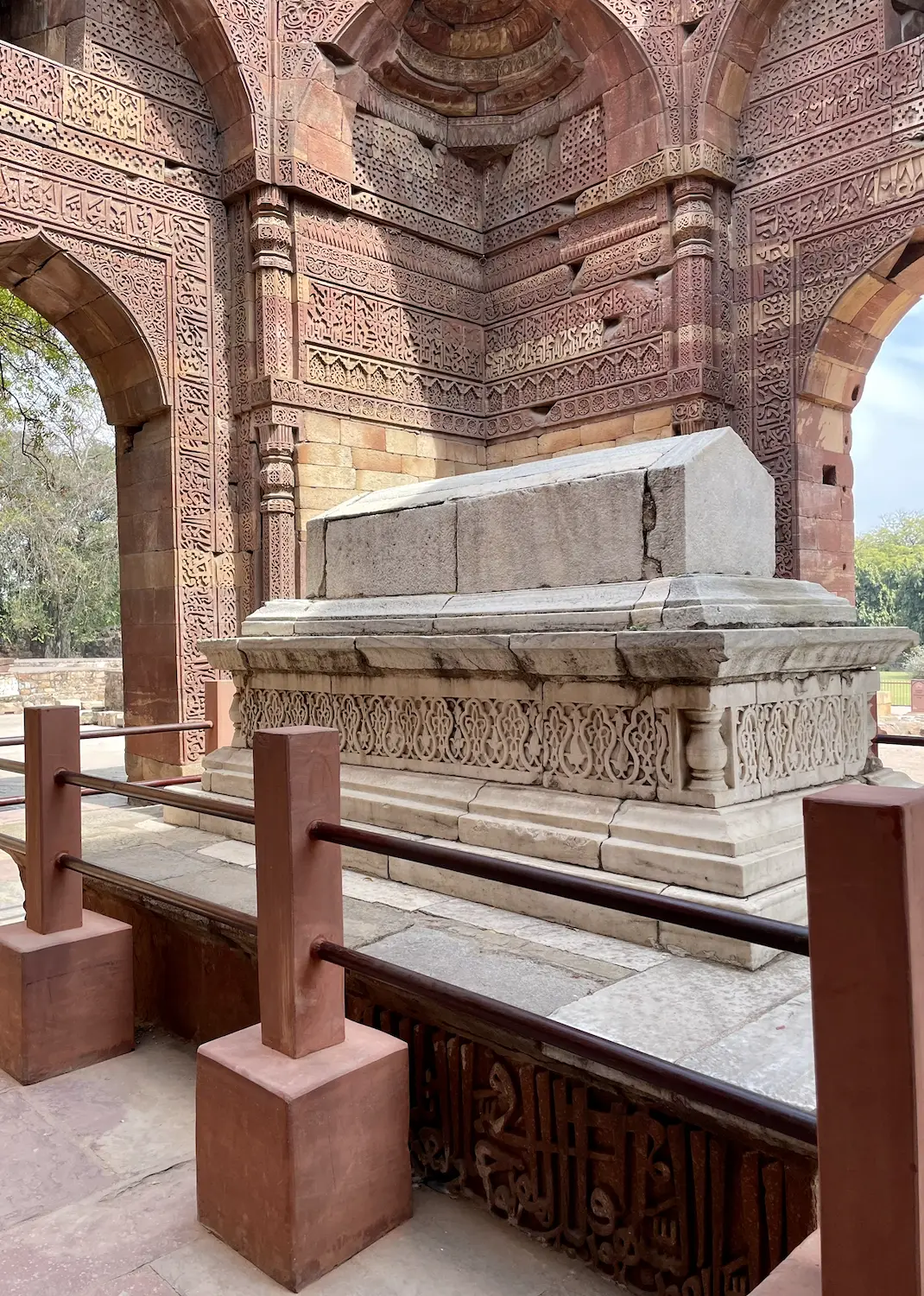 The white marble cenotaph of Iltutmish inside his tomb chamber, surrounded by densely carved red-sandstone walls with Arabic inscriptions and a low railing
