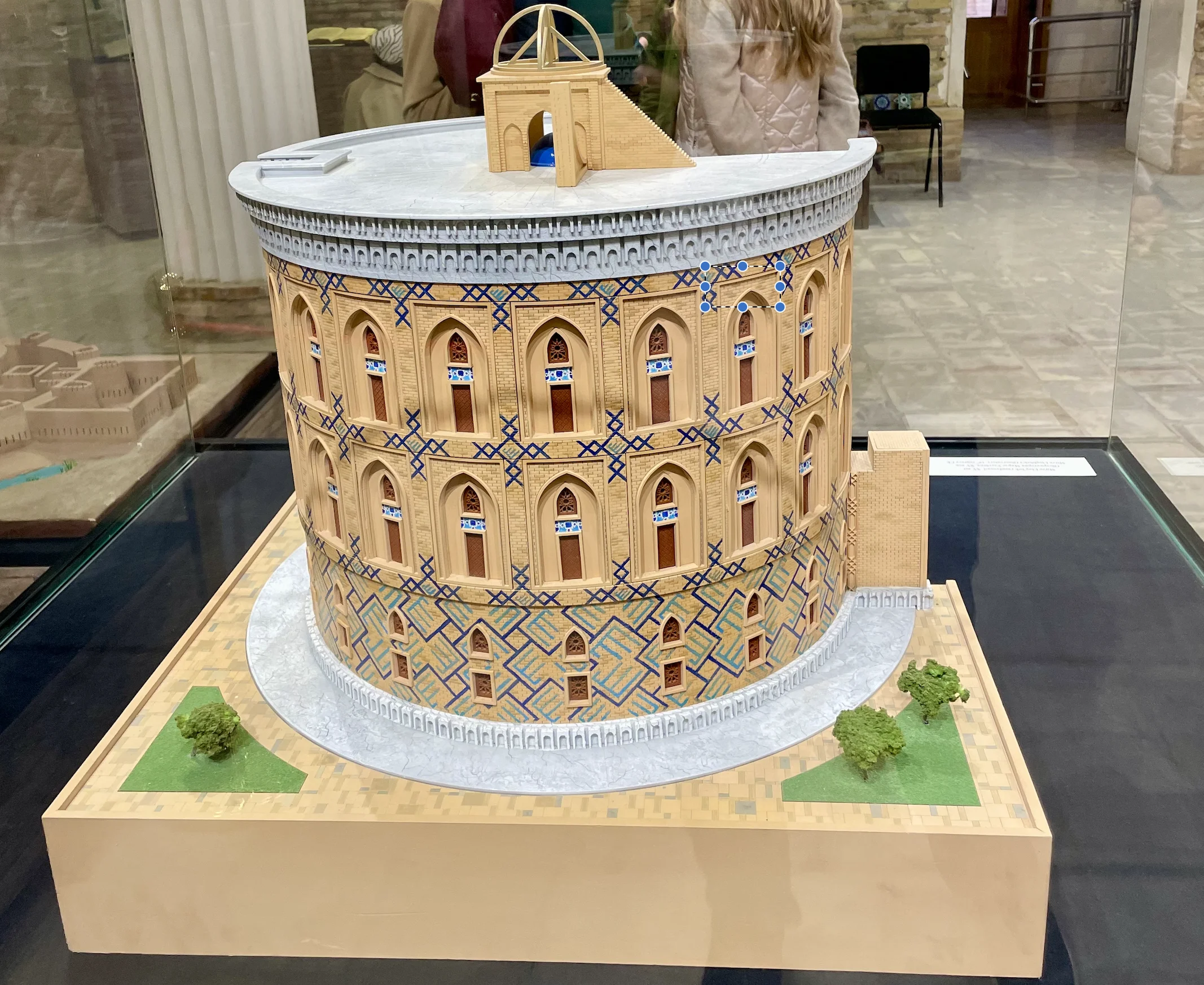 A scale model of a cylindrical building faced with blue and gold tile patterns and rows of arched niches, with the arc of a large instrument rising from its flat roof, displayed on a square base in a glass case, with museum visitors partly visible behind it