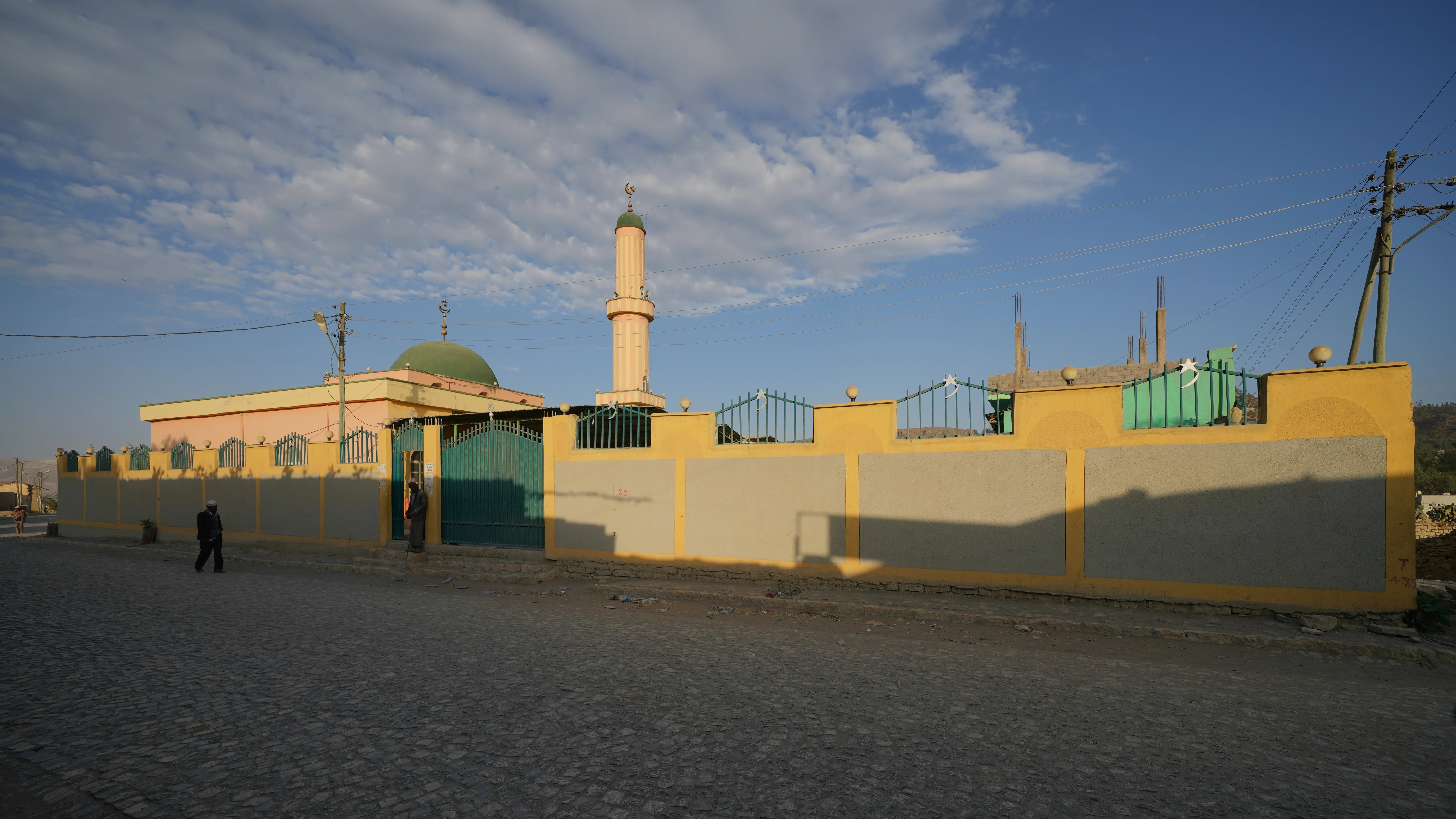 A walled mosque compound on a cobbled street in Mekelle, Ethiopia, with a pink and cream prayer hall under a green dome and a striped minaret topped by a crescent, evening light on its boundary wall, two men in the gateway and street traffic in the foreground
