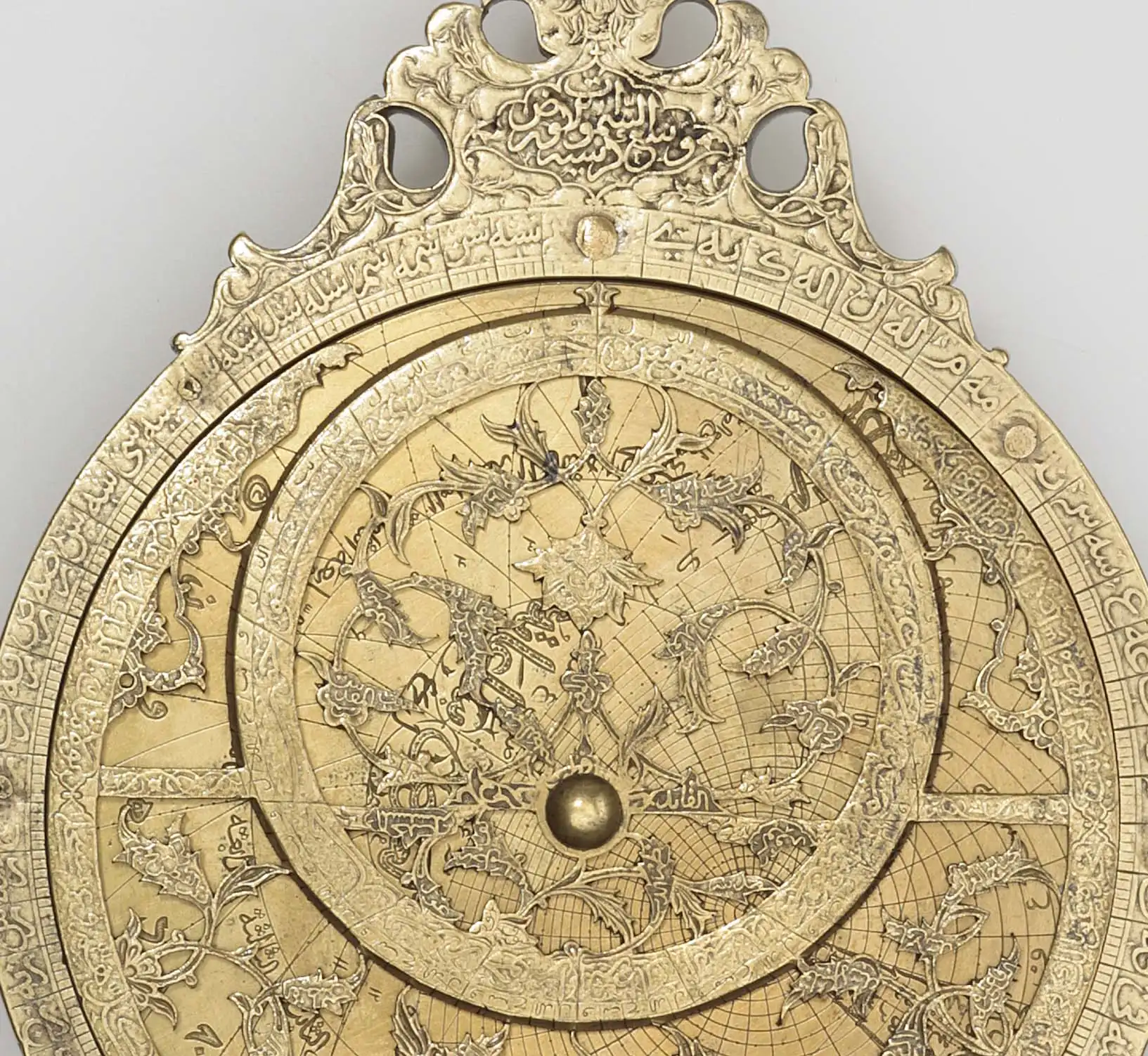 A brass-coloured astrolabe on a plain grey background, with a pierced cartouche of scrollwork at the top, a rim engraved with a degree scale and Arabic letters, and a pierced rete with star pointers over an engraved plate